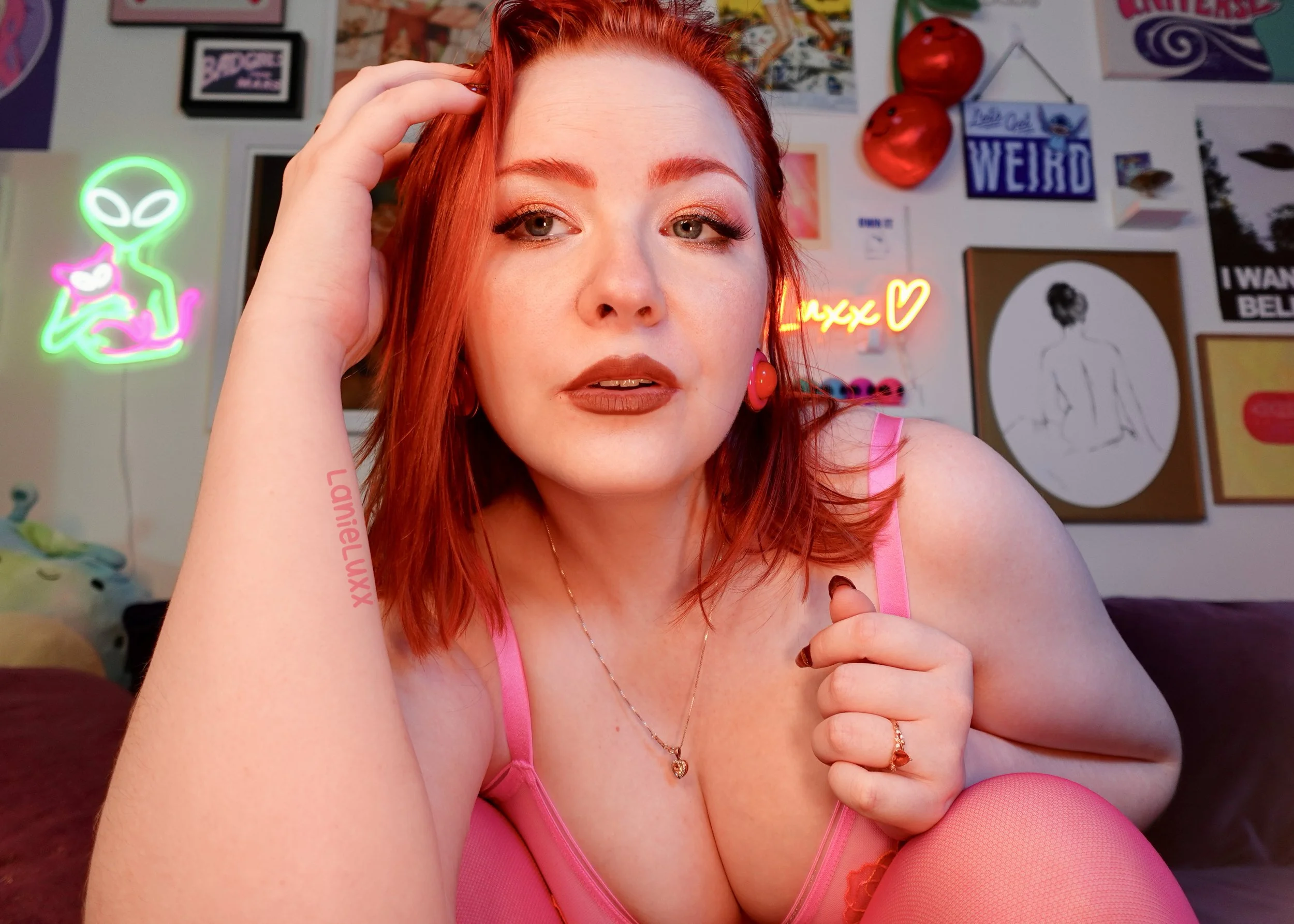 A woman with red hair and makeup wearing a pink top and fishnet stockings, sitting in a room decorated with neon and art posters