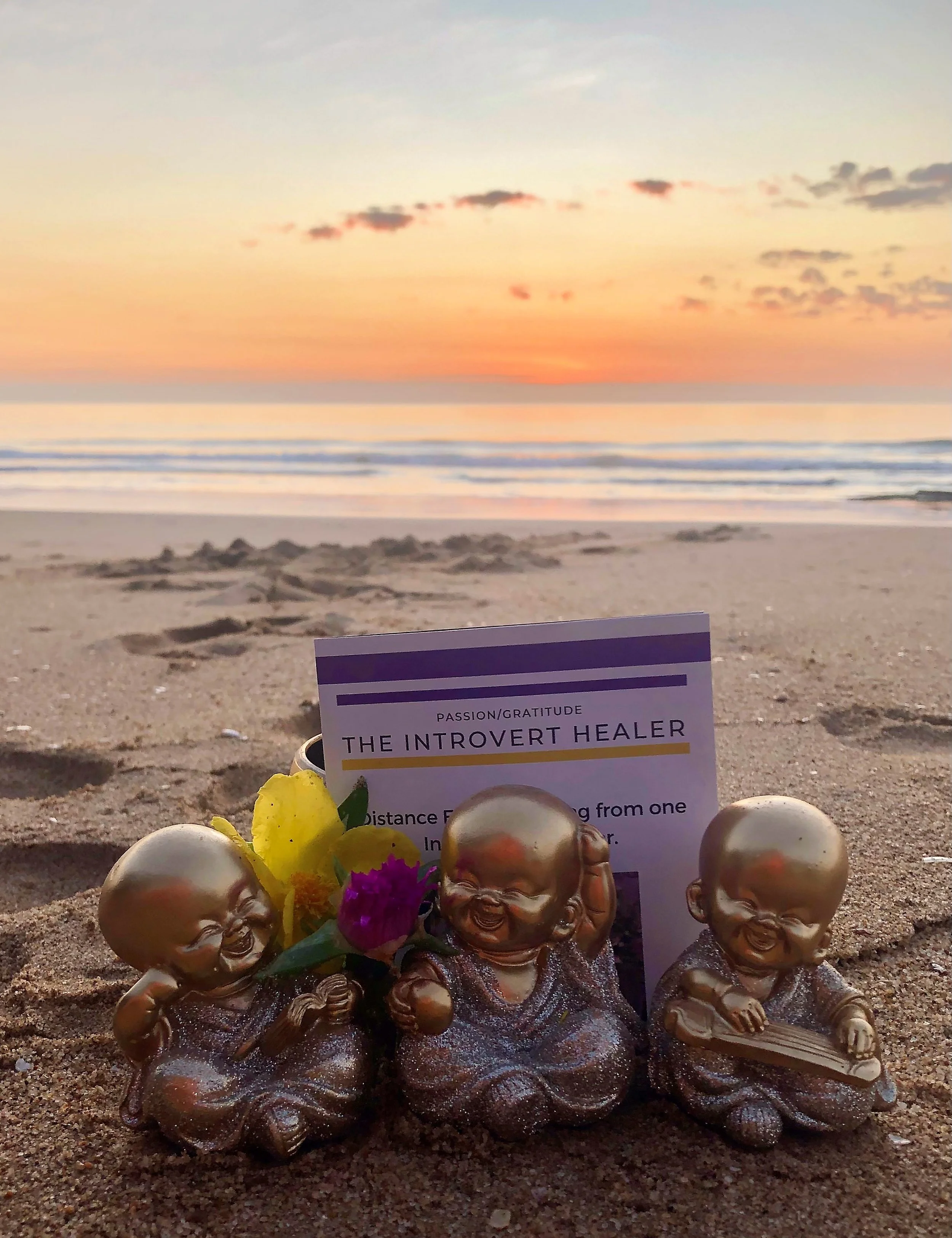 Three metallic Laughing Buddha figurines sitting on the sand at sunset, with flowers and a card behind them, on a beach.
