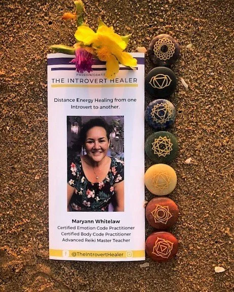 The Introvert Healer pamphlet in the sand. Above the pamphlet are one purple flower and one yellow flower. To the left are chakra stones