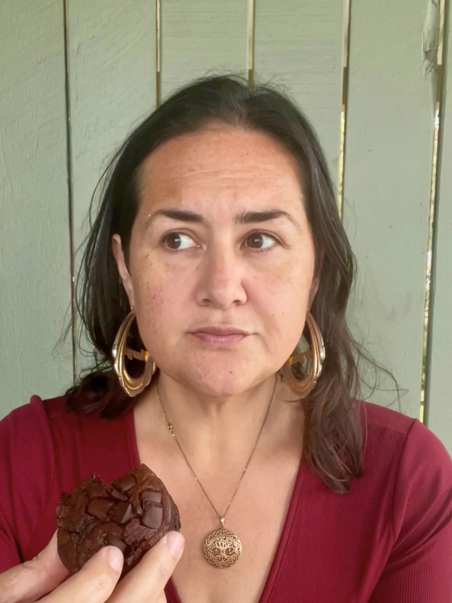 Brown haired woman, holding a chocolate muffin, wearing gold earrings and maroon shirt. Looking to side with contemplative glance.