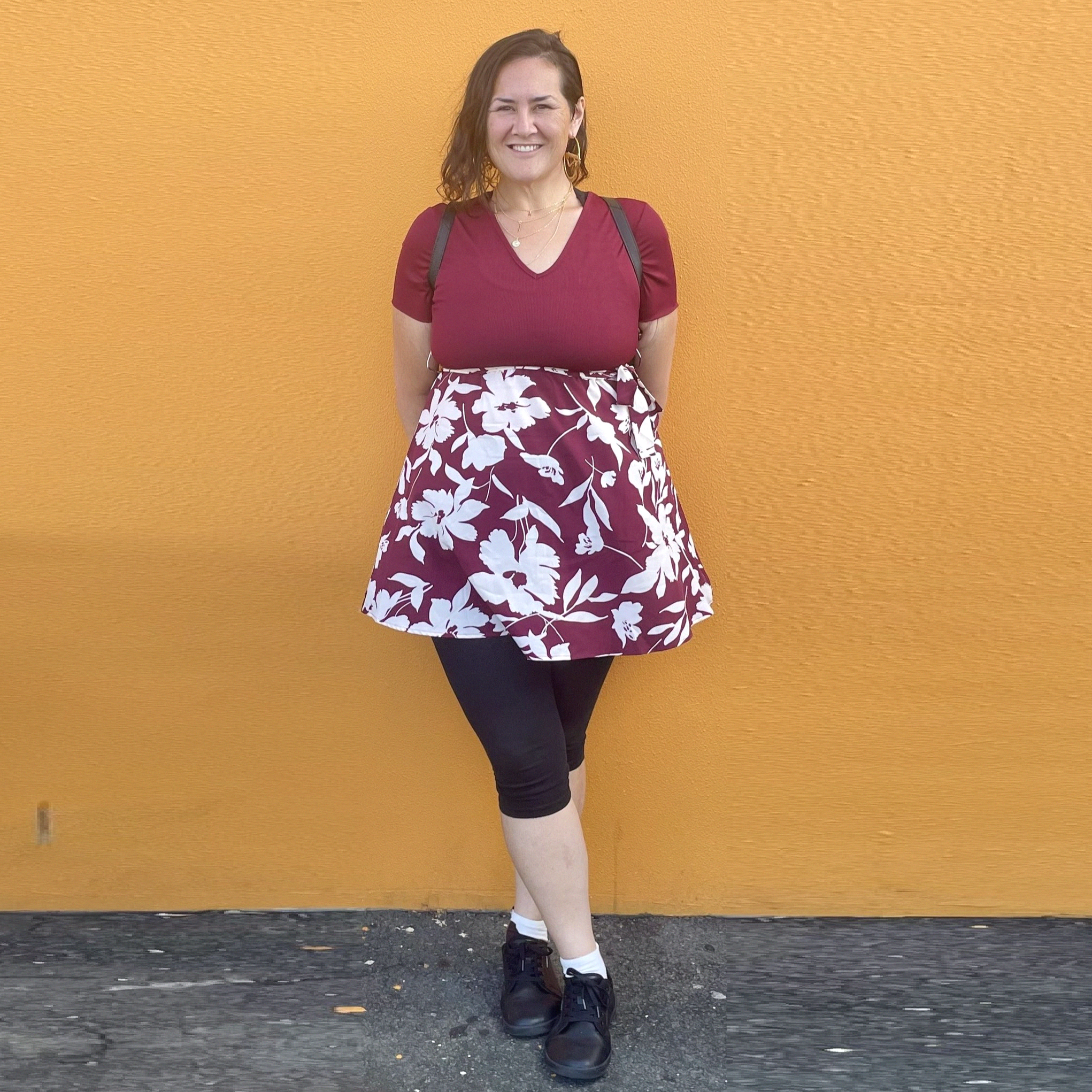 A woman with shoulder-length brown hair standing in front of an orange wall, smiling, wearing a maroon top, a floral skirt, black leggings, and black sneakers.