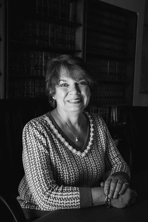 A woman with short hair smiling at the camera, sitting at a table with bookshelves in the background, in black and white.