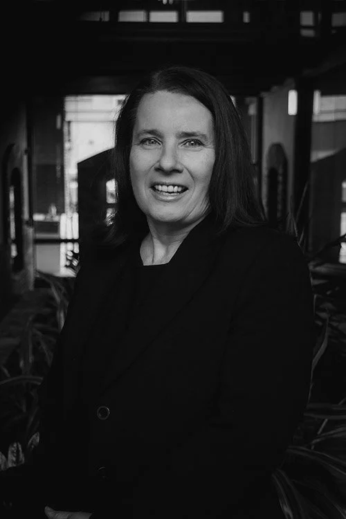 A black and white photo of a woman with shoulder-length hair, smiling, in an indoor setting with large windows and plants in the background.
