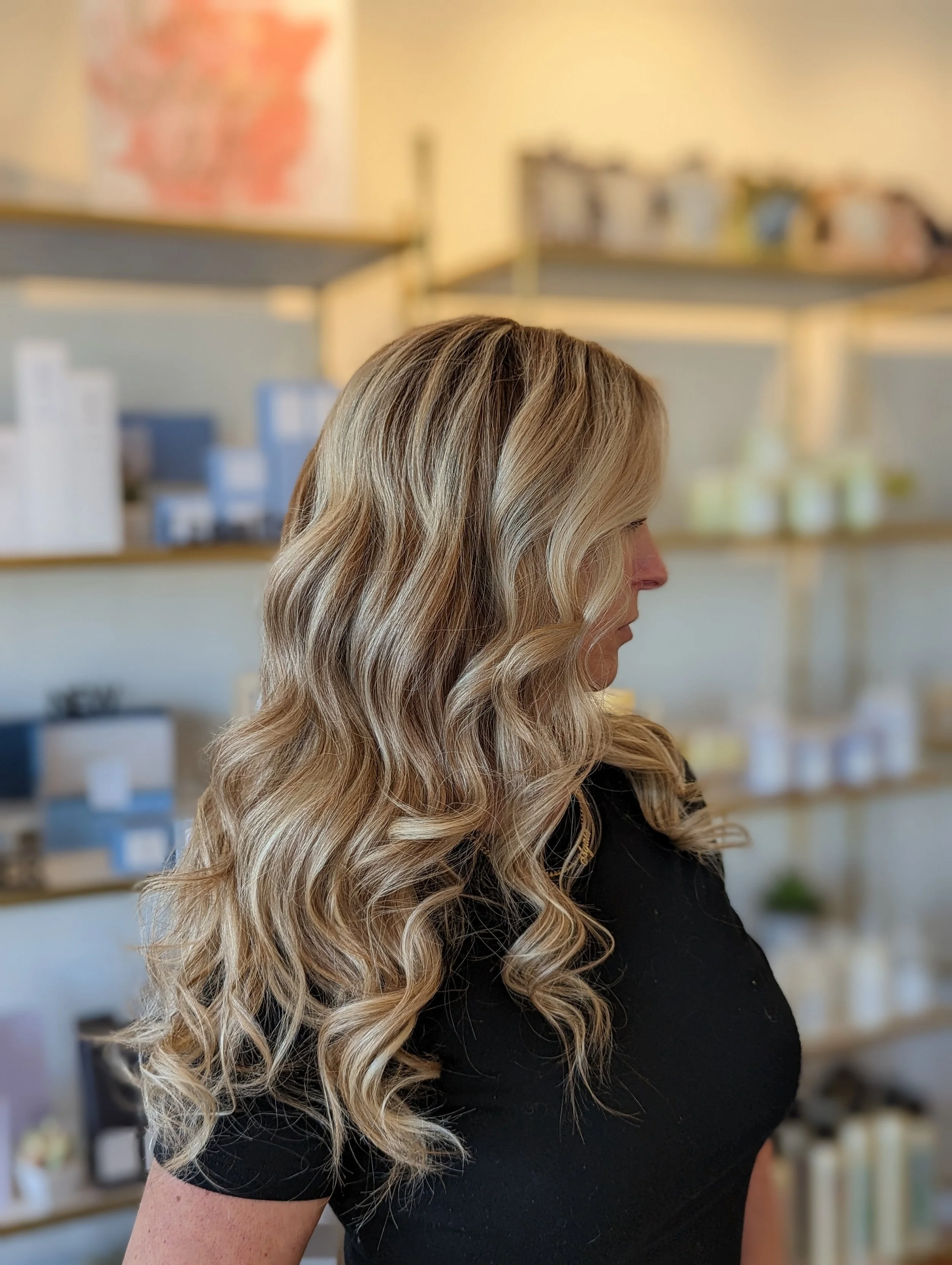 Side profile of a woman with long, blonde, wavy hair wearing a black shirt, standing in front of shelves with various products.