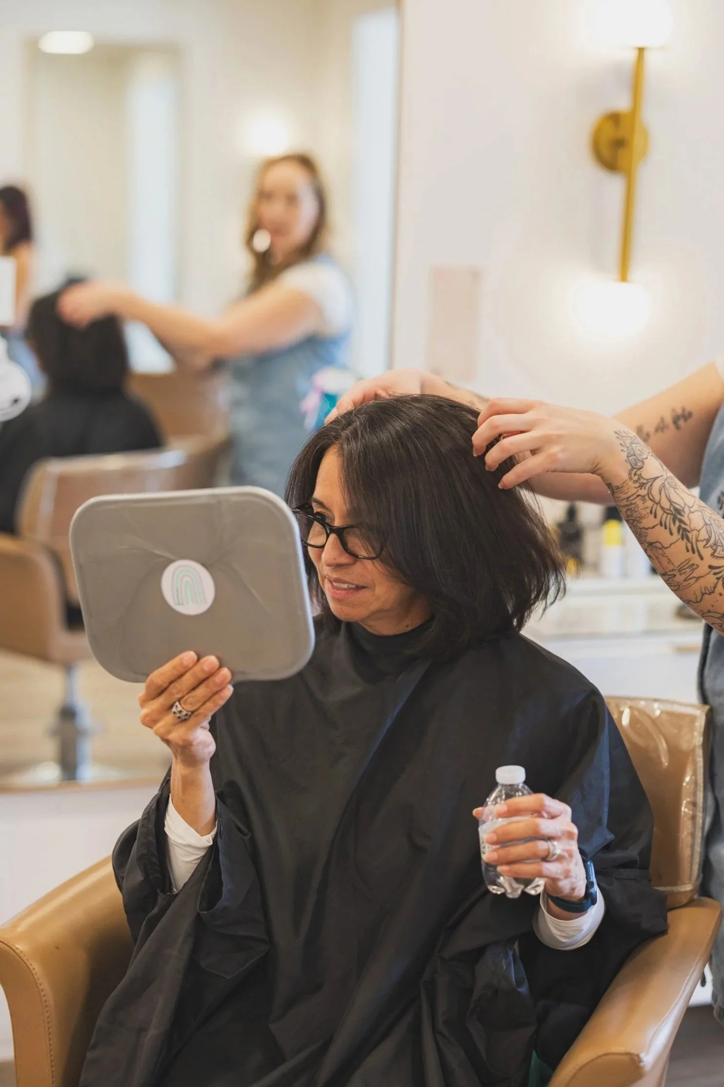 A woman with dark hair and glasses looks at herself in a mirror while a hairstylist styles her hair in a salon. The woman holds a water bottle, and another person is working on a client's hair in the background.