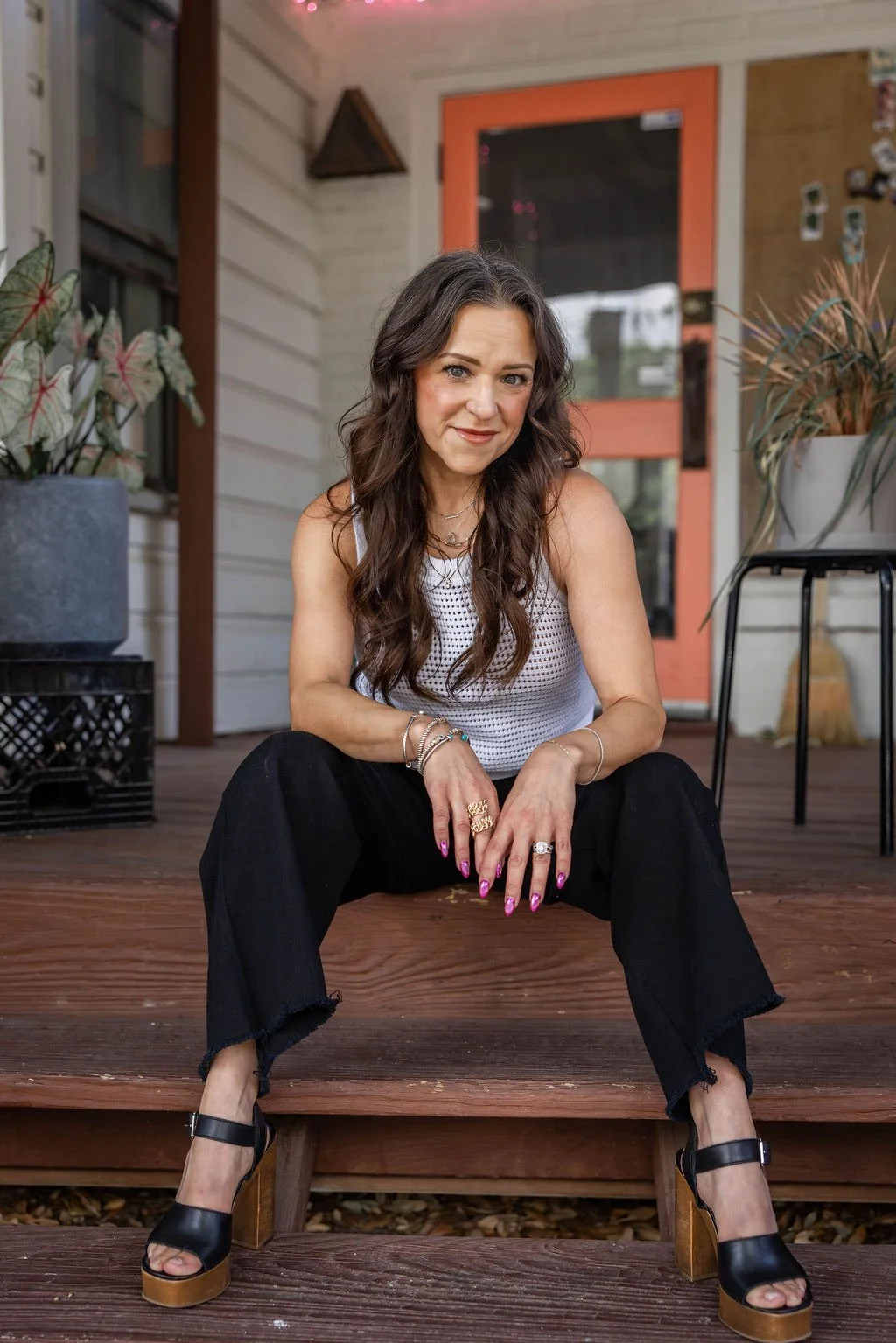 Woman sitting on porch steps with plants and a door in the background.