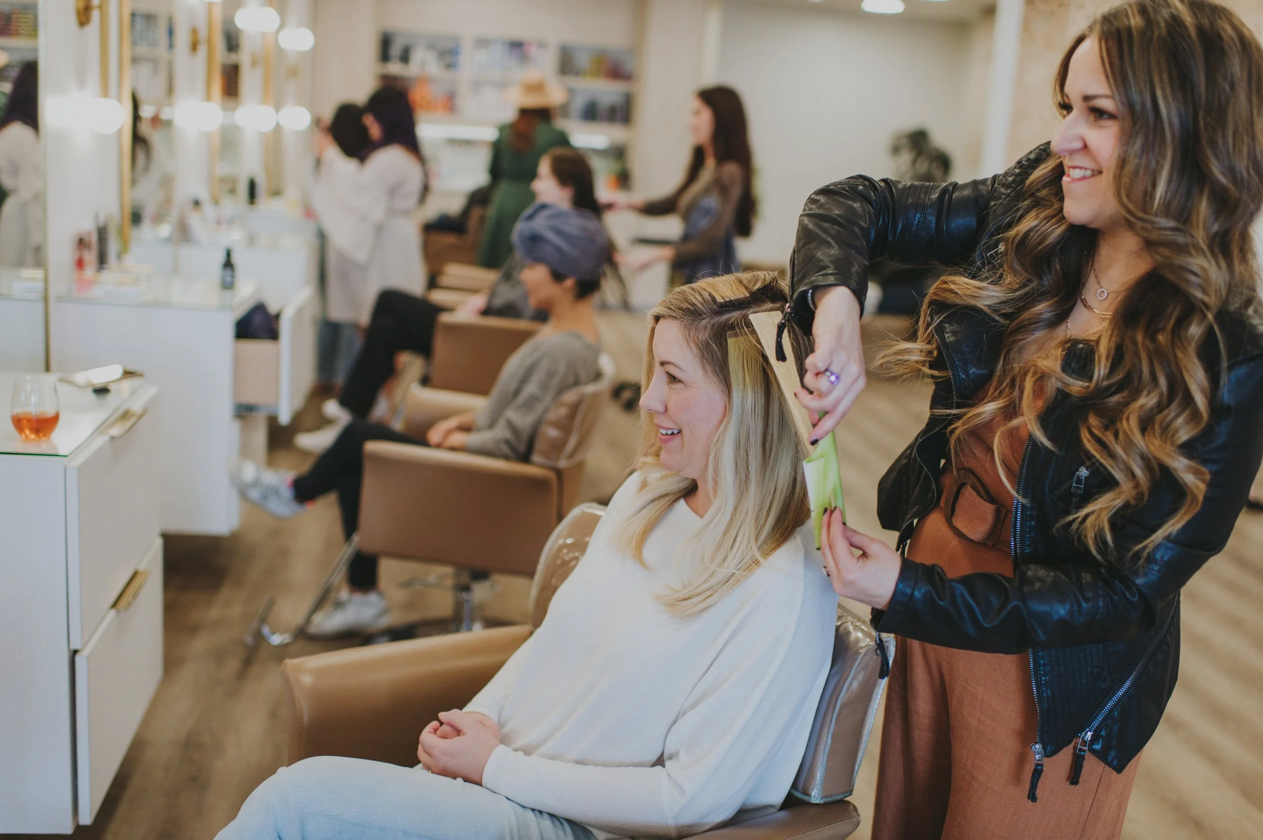 A woman getting her hair styled at a salon with other clients seated in the background.