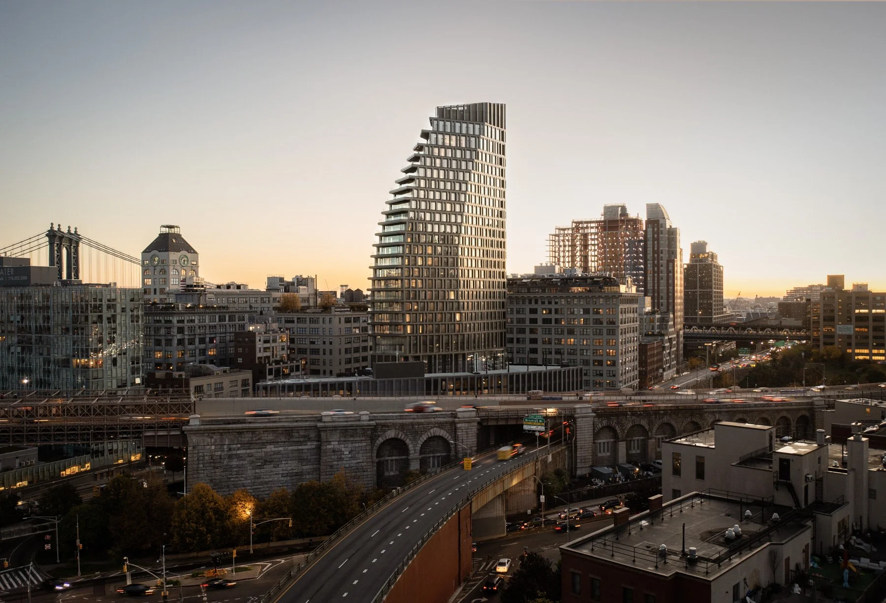 The tower rises above the surrounding DUMBO neighborhood at golden hour, with the BQE and Brooklyn skyline spread out behind it. The warm glow of lit interiors against the fading sunset sky makes the building feel alive and inhabited.