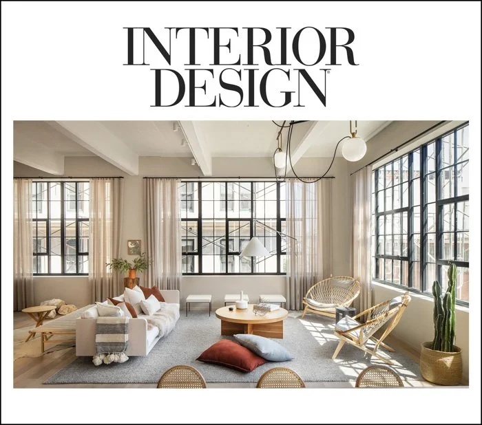 A modern living room with large grid windows, white walls, ceiling beams, and industrial-style light fixtures. The room features a white sofa, various pillows, a round wooden coffee table, wicker chairs, a cactus plant, and sunlight streaming through the windows.