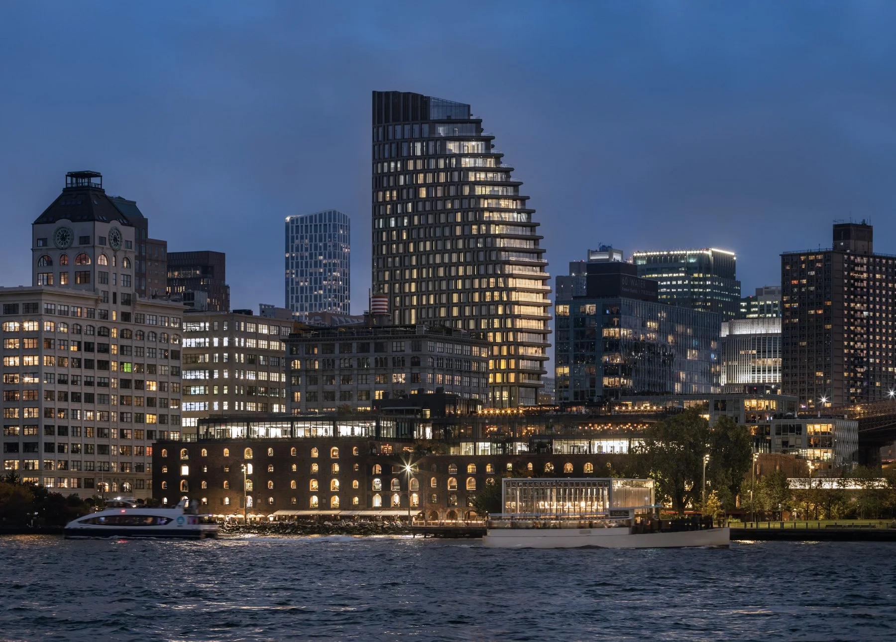 Photographed from across the East River at blue hour, the tower stands tall amid the glowing DUMBO waterfront, with the historic Empire Stores and Jane's Carousel pavilion in the foreground.