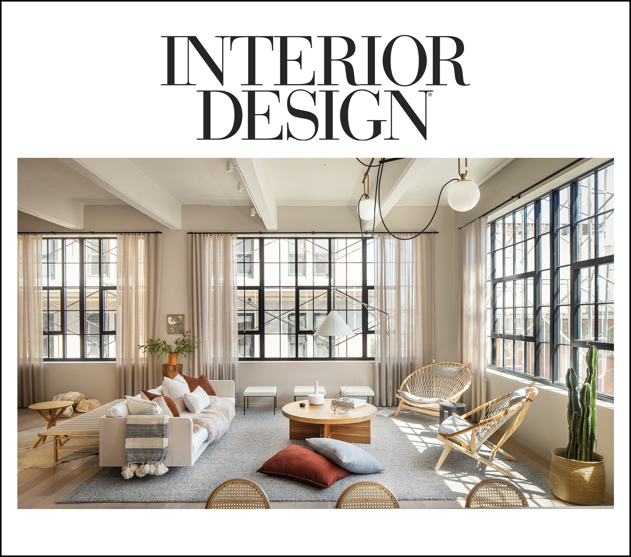 A modern living room with large grid windows, white walls, ceiling beams, and industrial-style light fixtures. The room features a white sofa, various pillows, a round wooden coffee table, wicker chairs, a cactus plant, and sunlight streaming through the windows.