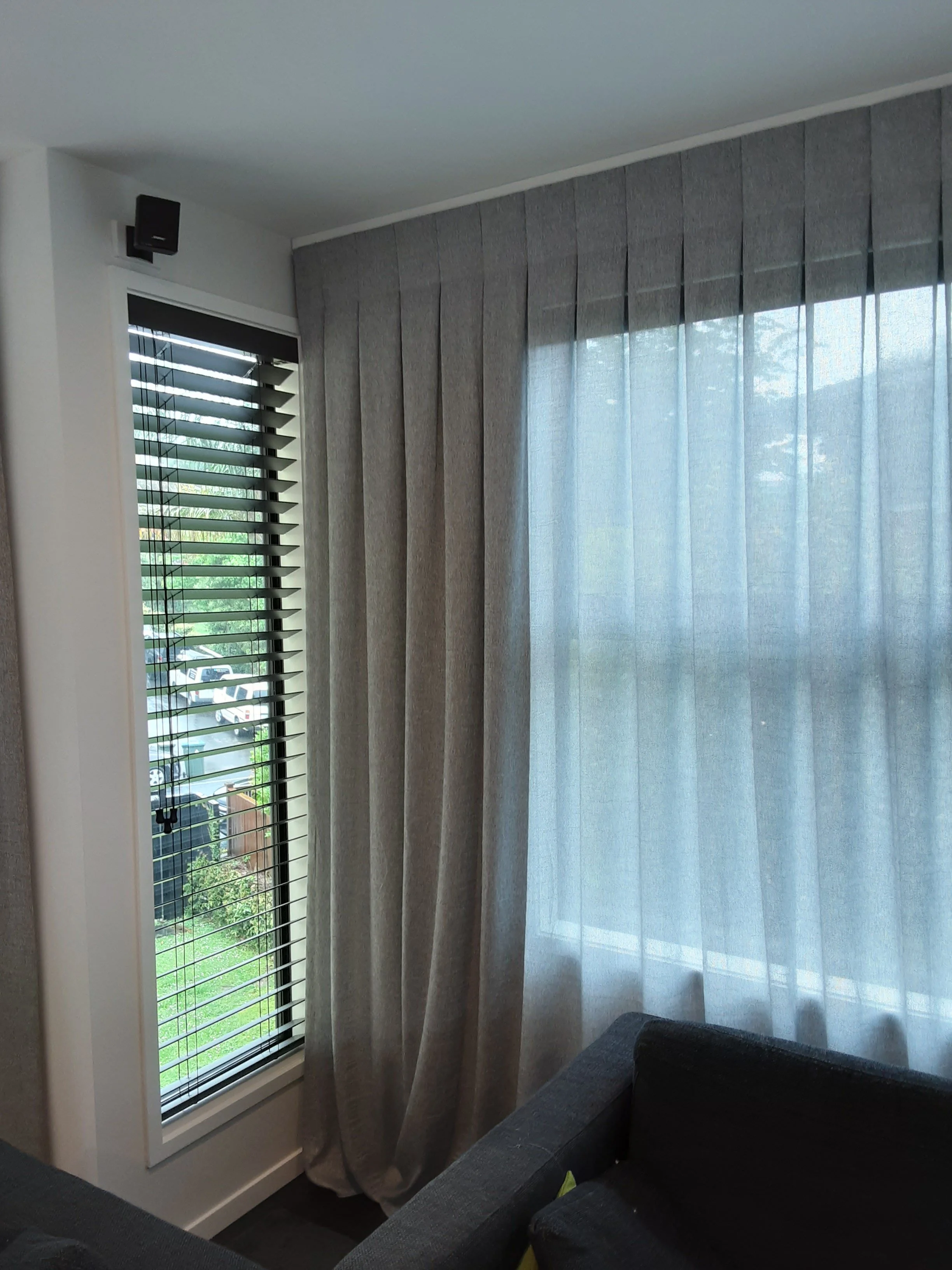 Interior view of a room corner with a window covered by vertical blinds and sheer reverse pleat (Inverted Pleat) curtains, a black sofa with a yellow cushion, and a small black wall-mounted speaker near the ceiling.