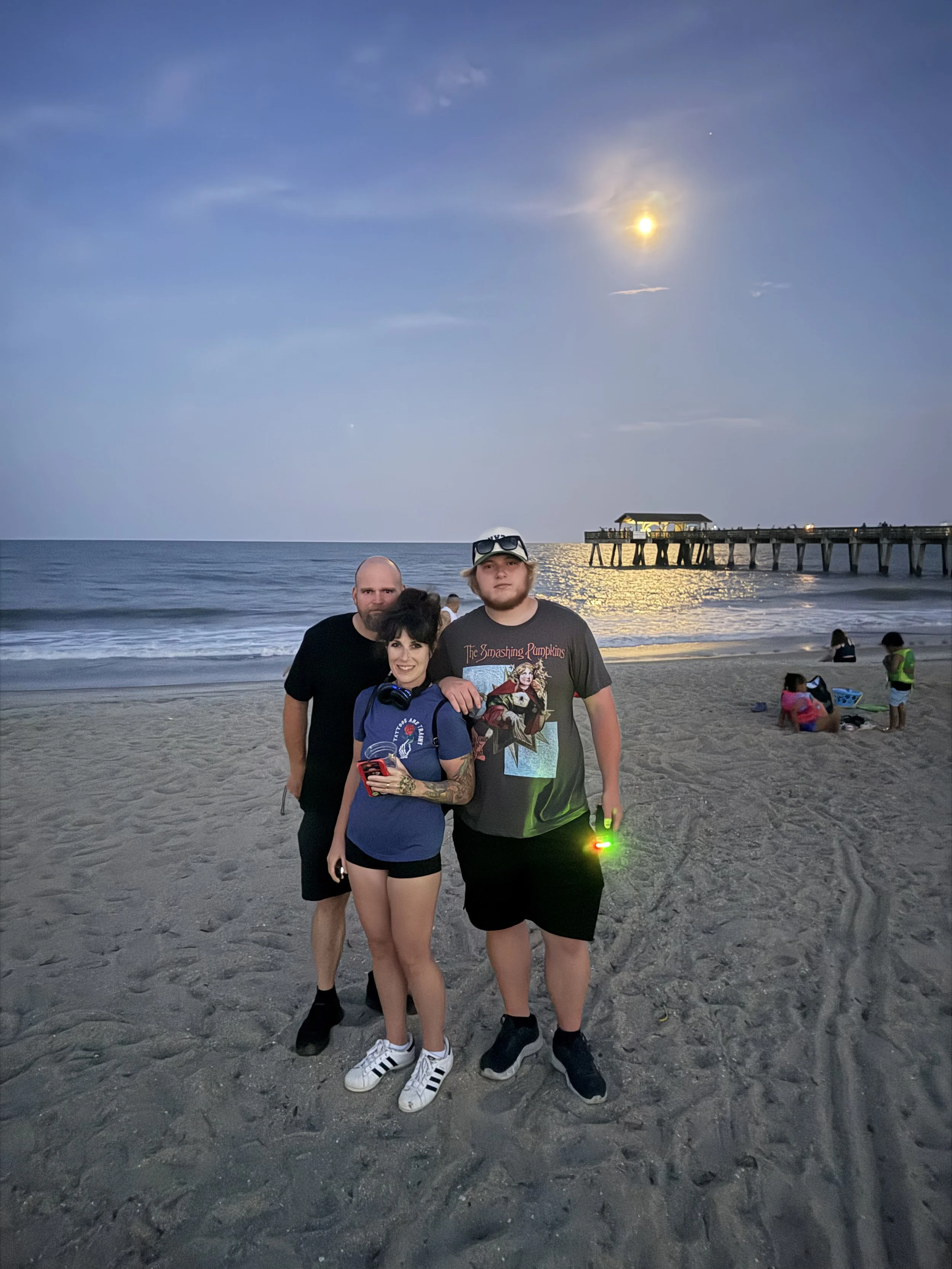 A group of four people on a beach during sunset or dusk, with a pier extending into the water in the background. The group includes two men, one woman, and a young man. They are standing close together, smiling and posing for the photo. The woman is wearing headphones around her neck, and the young man is holding a small glowing device in his hand. There are other people and children in the background enjoying the beach.