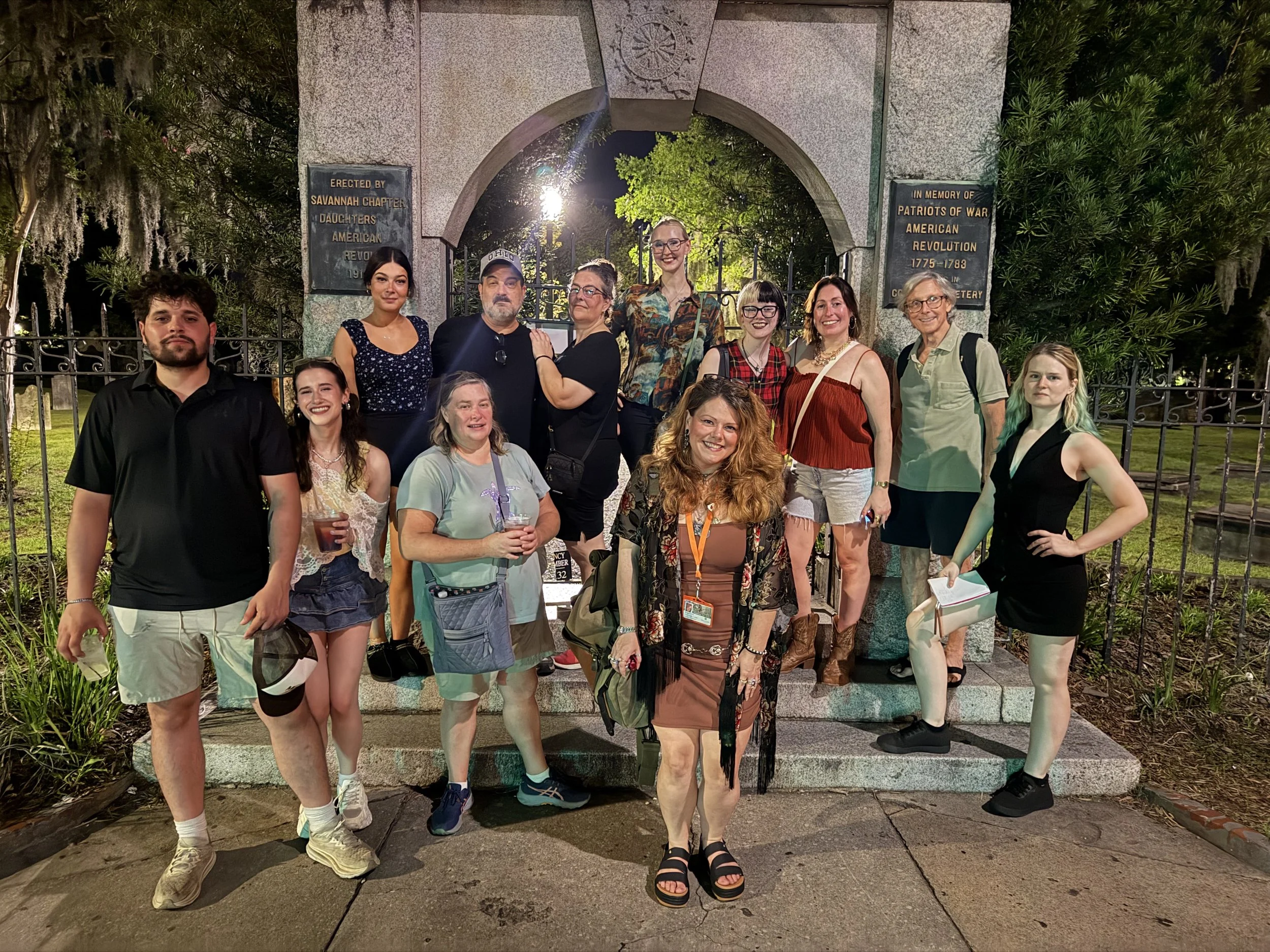 Group of twelve people standing and sitting in front of an archway at night, with trees and plaques nearby. They are smiling, some holding drinks, and dressed casually.