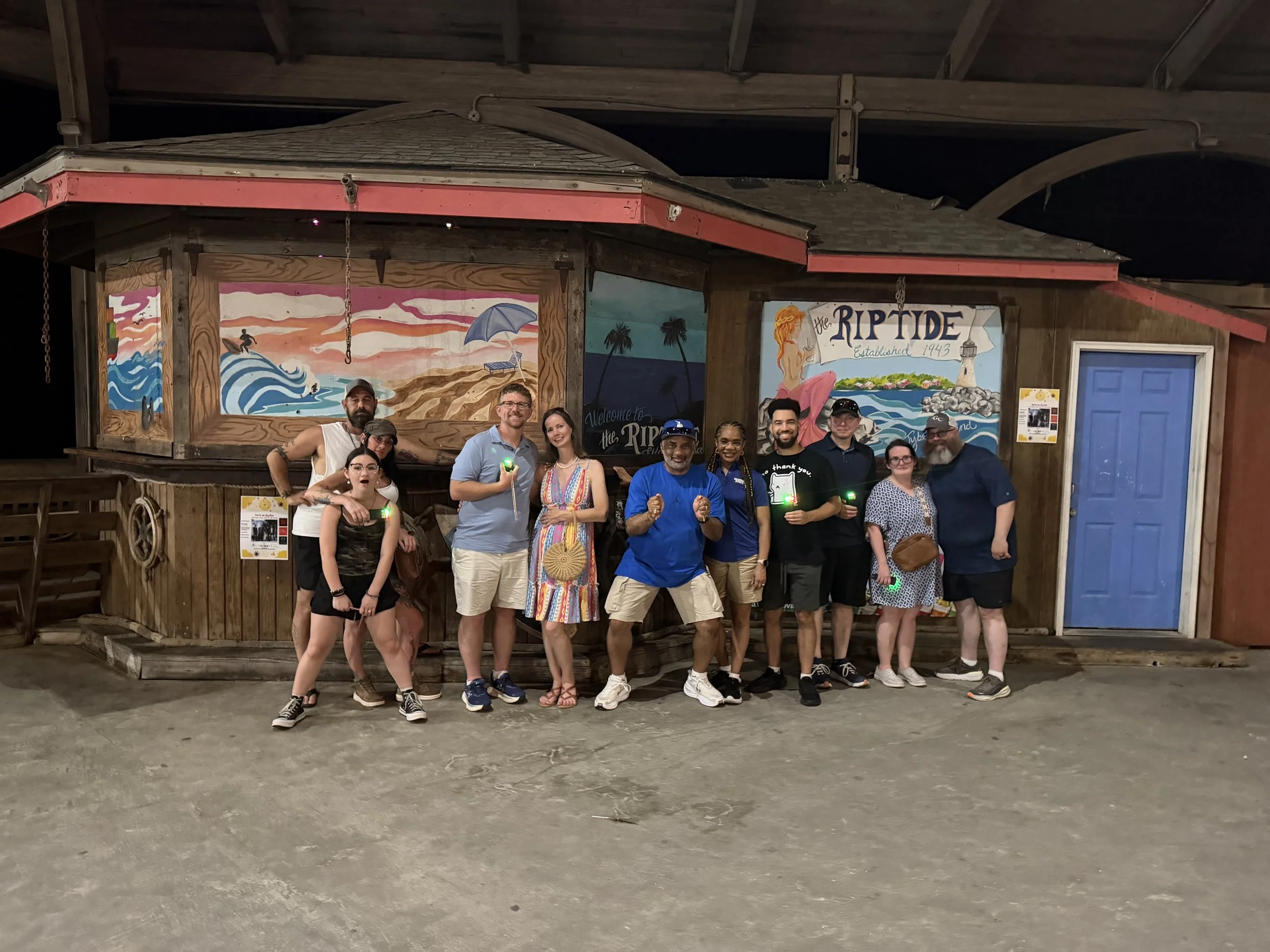 Group of people standing in front of a painted mural on a wooden building. The mural features ocean waves, a surfer, a beach umbrella, and a lighthouse, with the text 'The RIPTIDE Established 1943.' The group is smiling and some are holding glow sticks.