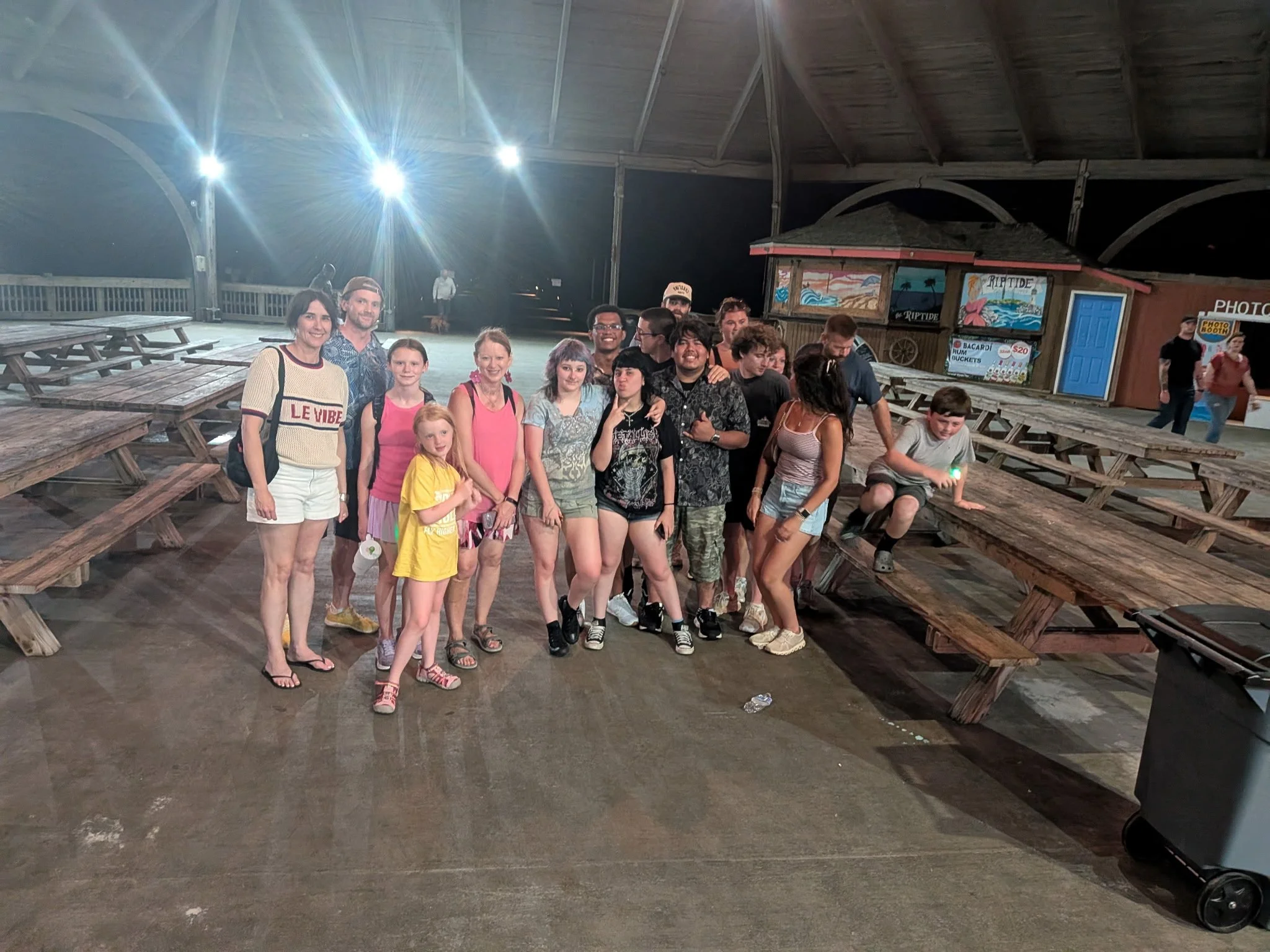 Group of people standing on a wooden pier at night, with picnic tables, a small building with a sign that reads 'Riptide,' and bright lights overhead in a beach setting.