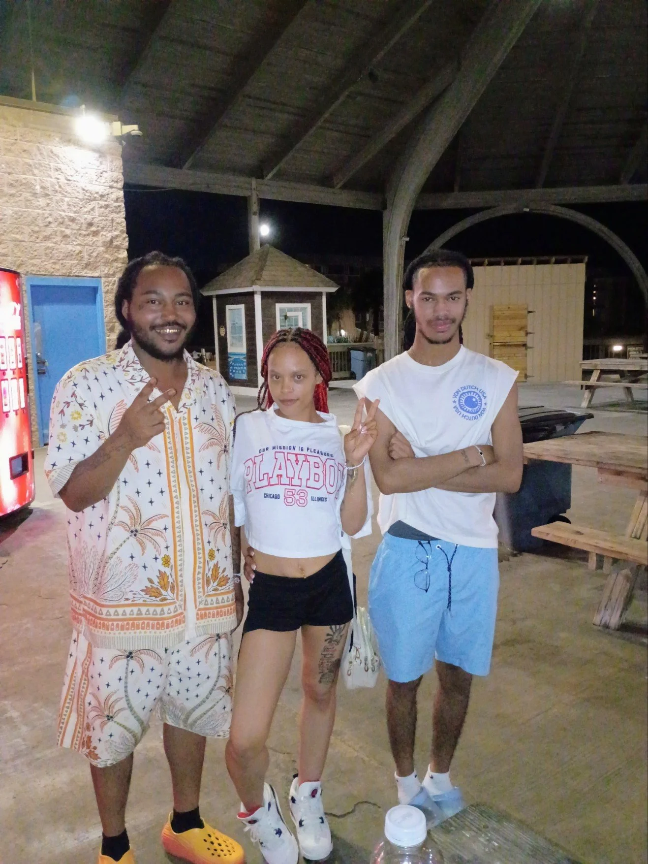Three men and one woman standing outside at night, smiling and making peace signs. They are casually dressed, with one wearing a patterned shirt and shorts, another in a Playboy T-shirt and shorts, and the young woman in a white T-shirt with black shorts. The background features a pavilion, picnic tables, and a vending machine.