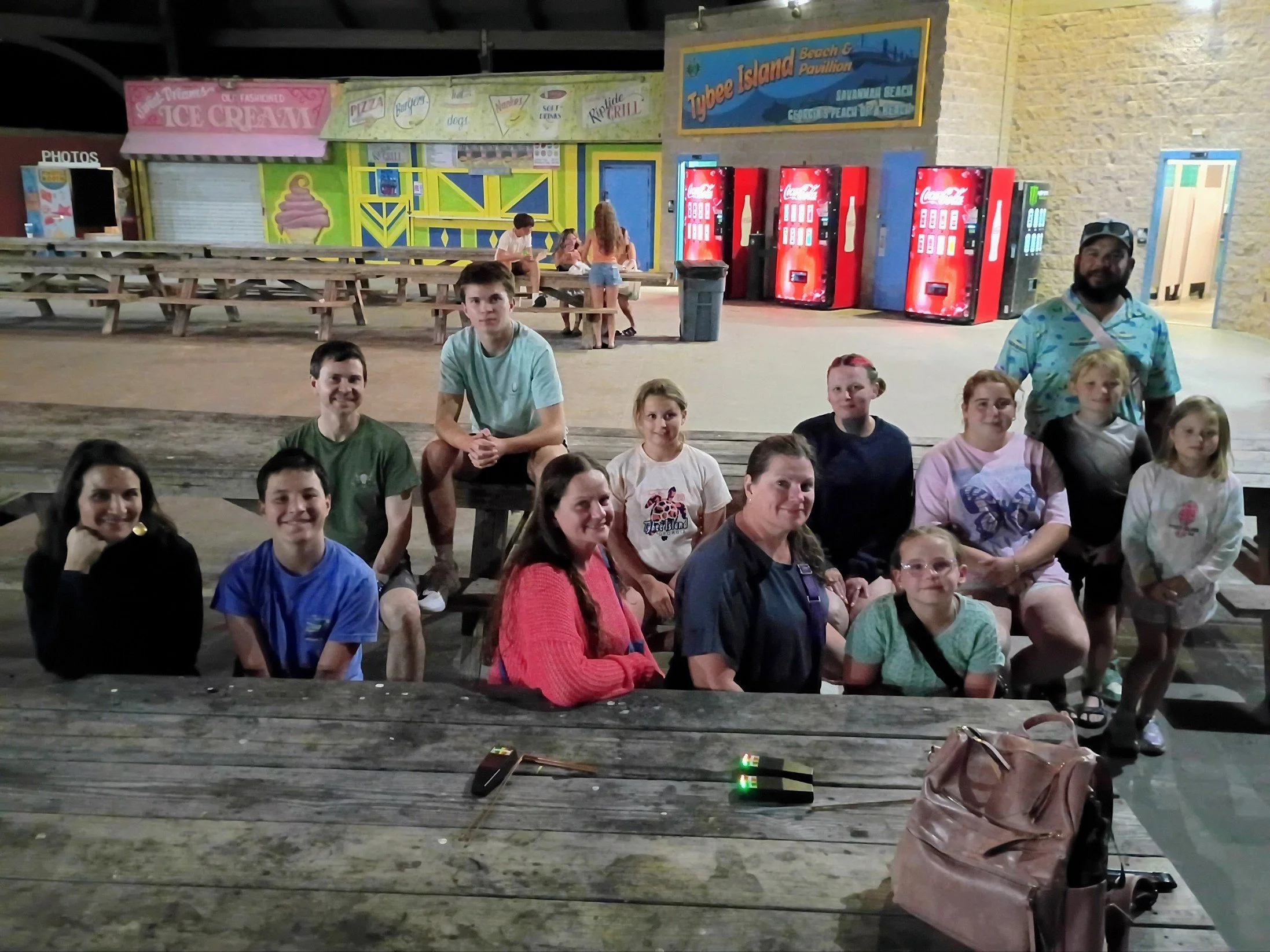 A group of people sitting on wooden picnic tables at Tybee Island Beach Pavilion at night, with a row of red Coca-Cola vending machines and a colorful amusement area in the background.