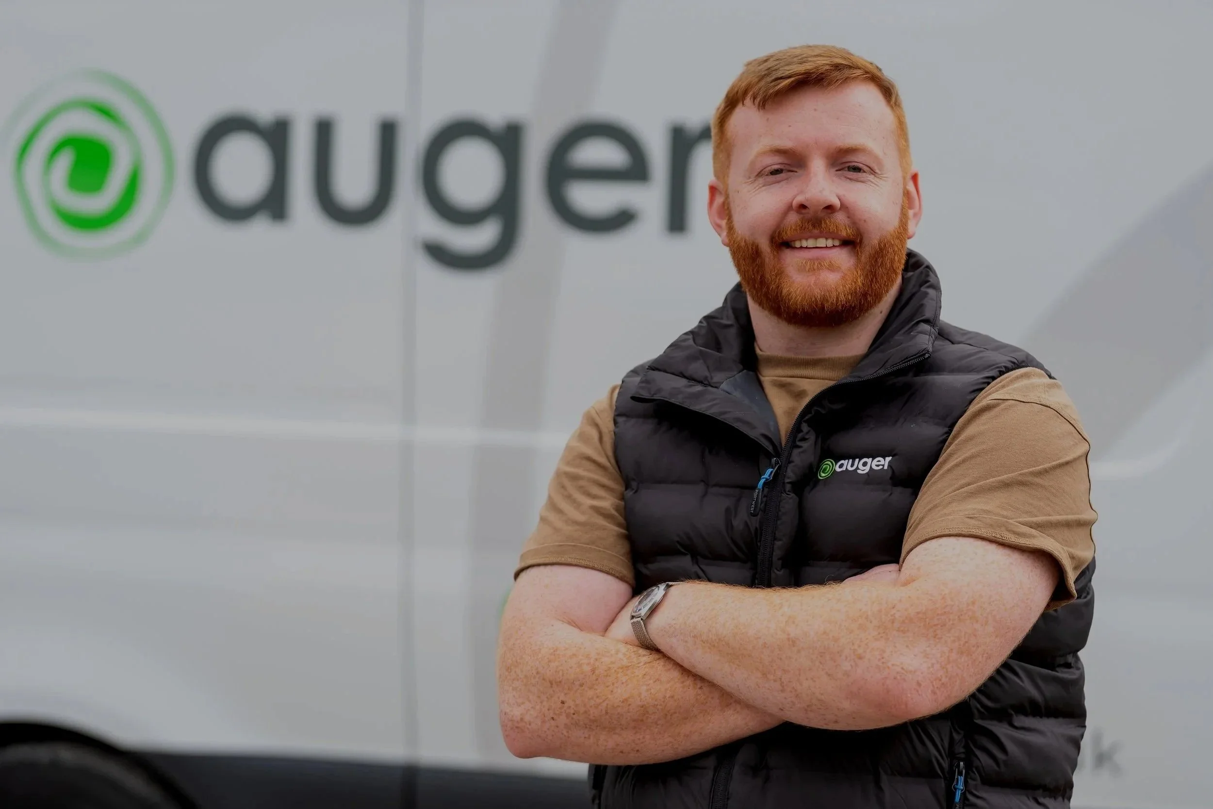 A smiling man with red hair and a beard standing outdoors in front of a vehicle with the 'Auger' logo on it, wearing a black vest over a tan shirt.