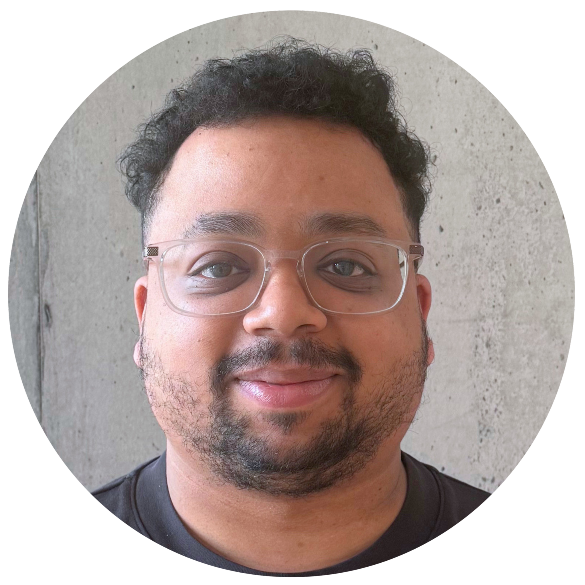 Close-up of a person's face wearing clear glasses, with short curly black hair, a slight smile, and facial hair, against a neutral gray background.