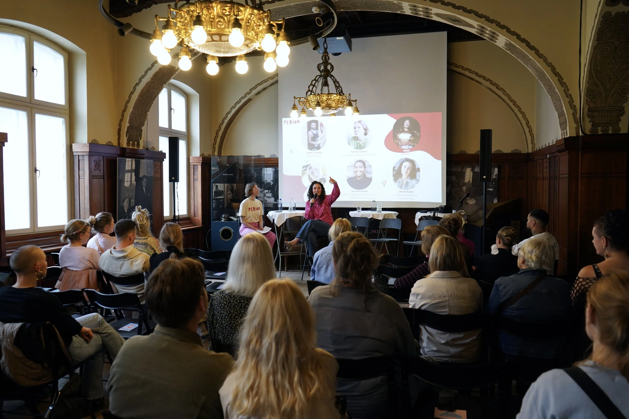 En gruppe unge mennesker står i en kontorindretning med flere computerskærme, mens to kvinder sidder foran computere og arbejder. Der er en skærm med teksten TV AVISEN 18:30.