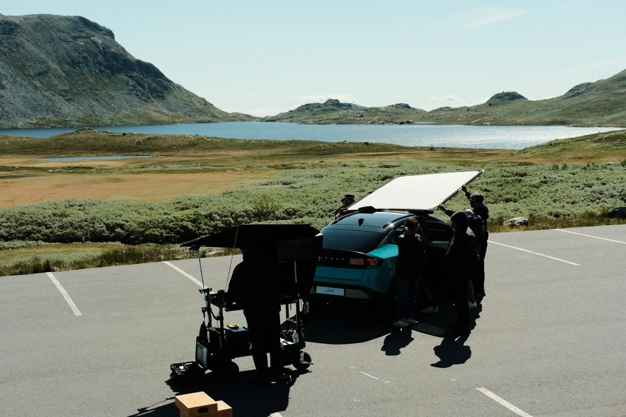 Behind the scene look at a film production in Norway. Group of people working around a teal electric car with its rear hatch open in a parking lot near a scenic lake and mountains.