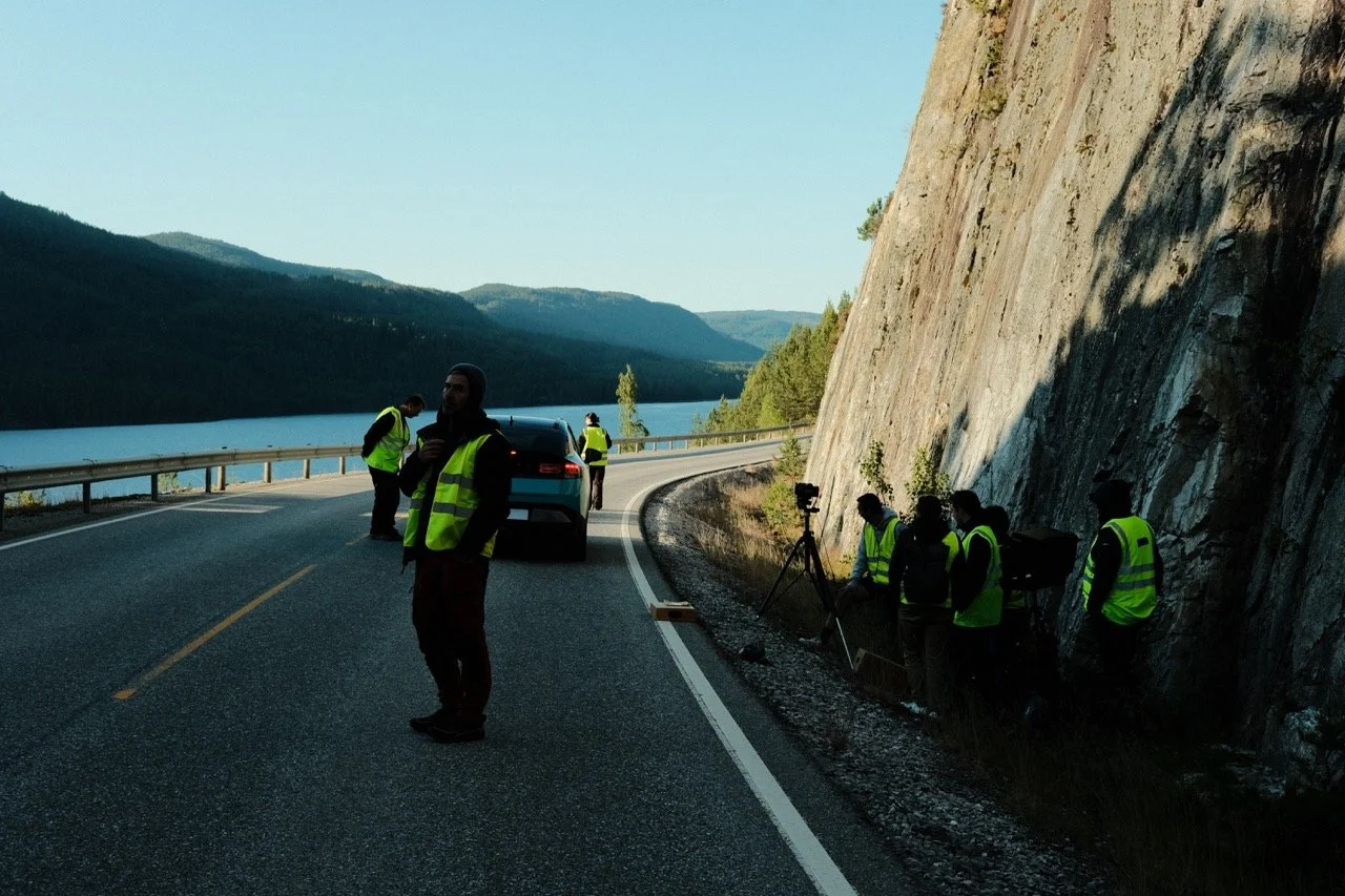 Group of people wearing yellow safety vests working on a road near a rocky hill and a body of water with mountains in the background.
