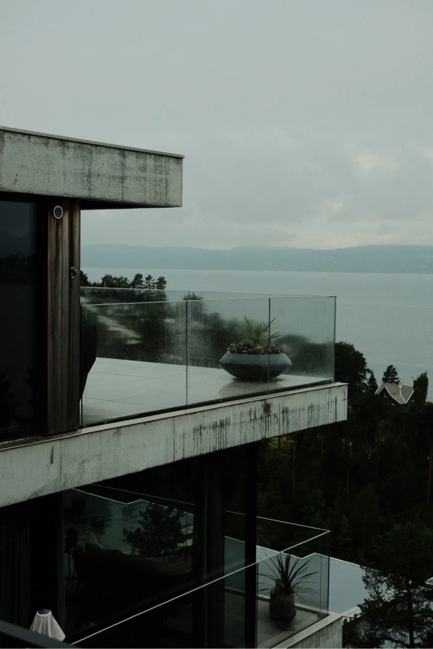 Behind the scene look at a film production in Norway. Monky Productions Production Service Norway. Modern apartment balcony with glass railing overlooking a body of water and distant hills, trees, and houses in the background on a cloudy day.