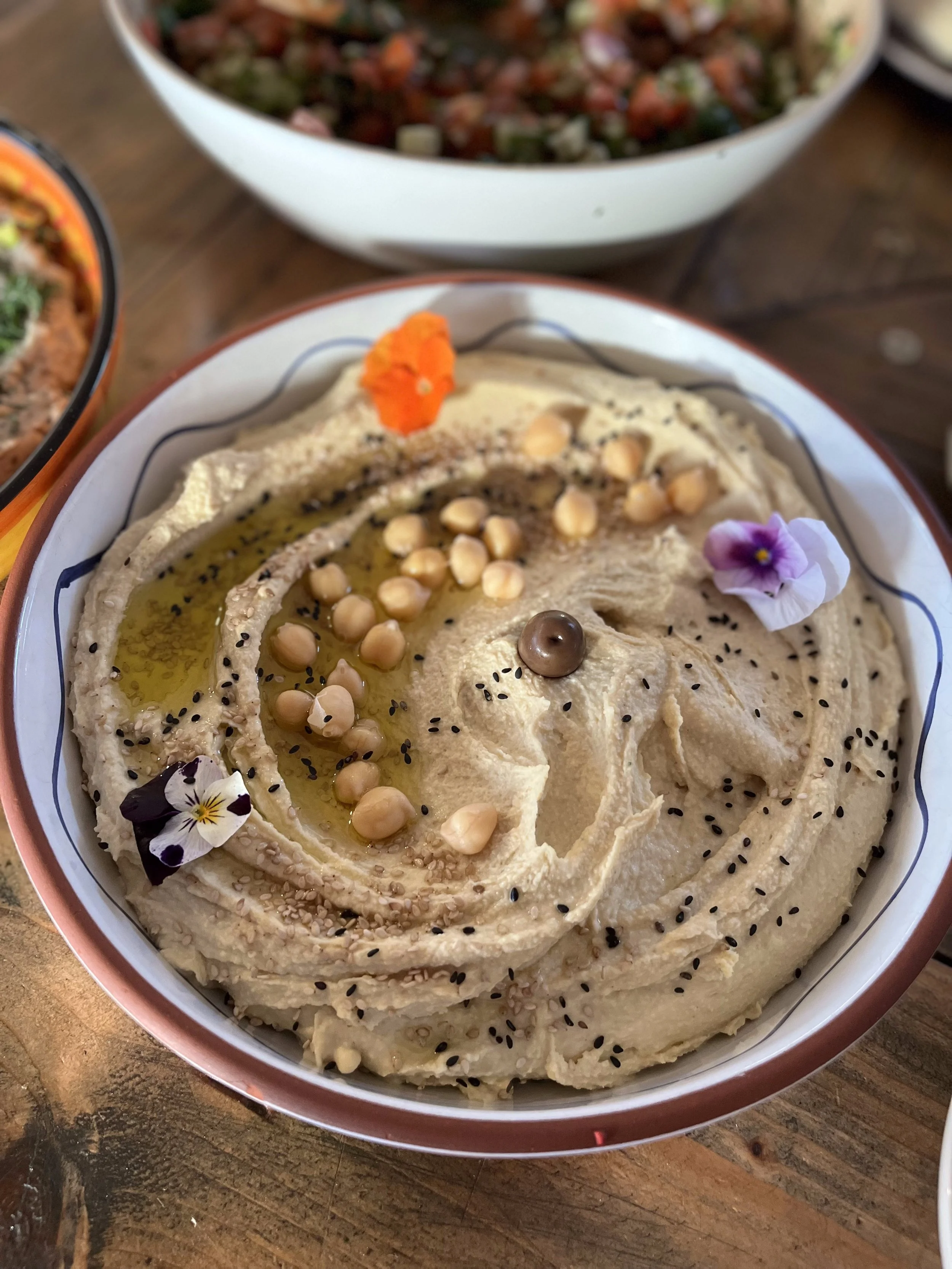 A bowl of hummus garnished with chickpeas, black sesame seeds, a single mushroom, paprika, black pepper, and edible flowers, drizzled with olive oil.