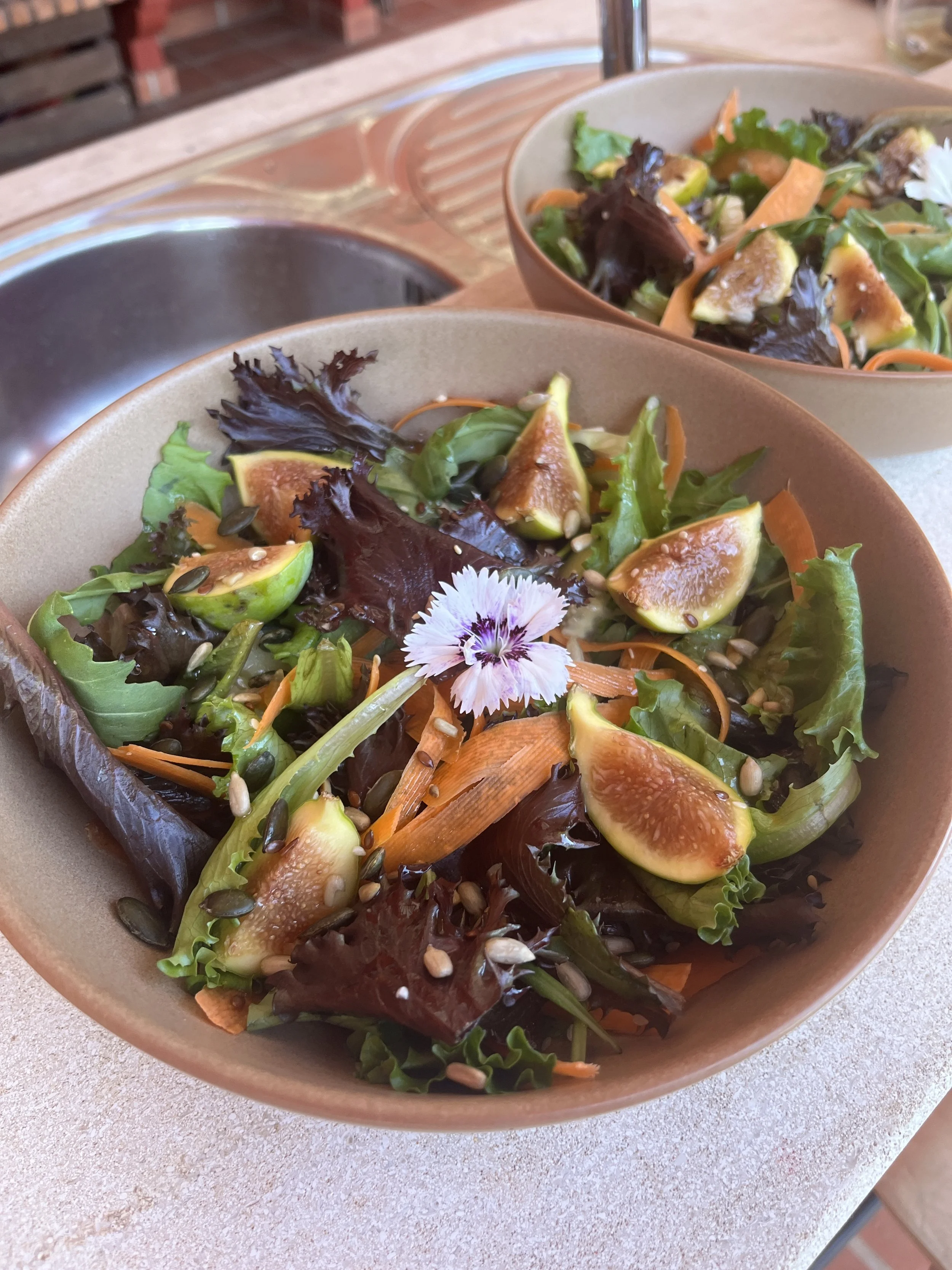 Bowl of mixed green and purple lettuce salad with figs, shredded carrots, sunflower seeds, and a flower garnish.