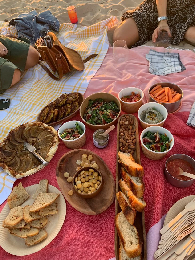 A picnic spread on a blanket with various dishes including bread, salads, hummus, carrots, nuts, and dips, with people sitting nearby on a beach.