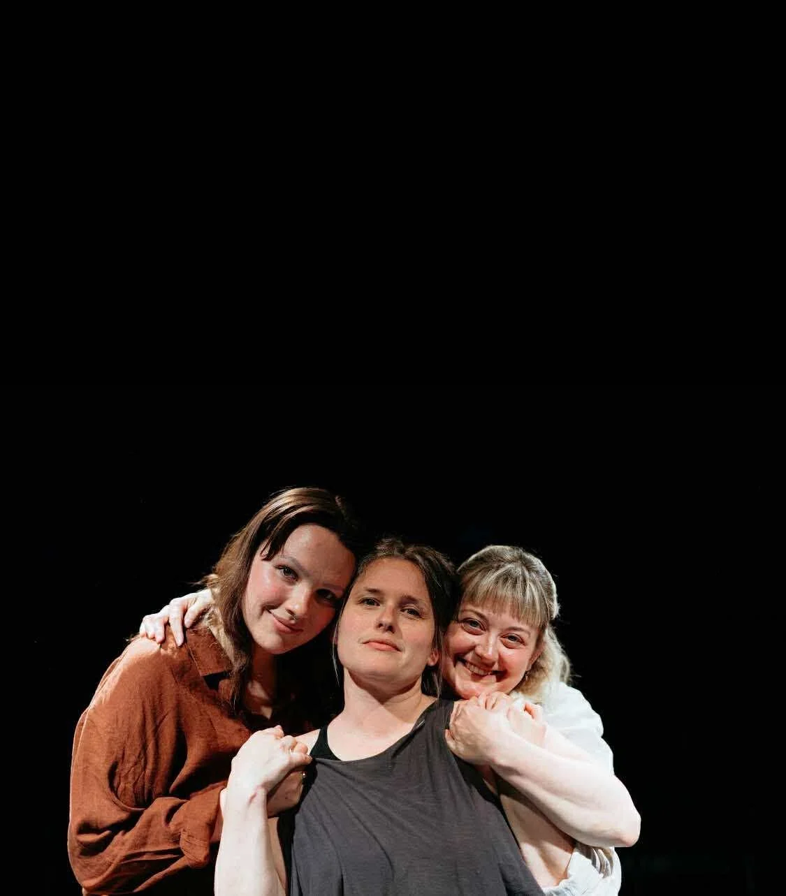 Three women embracing and looking at the camera against a black background.