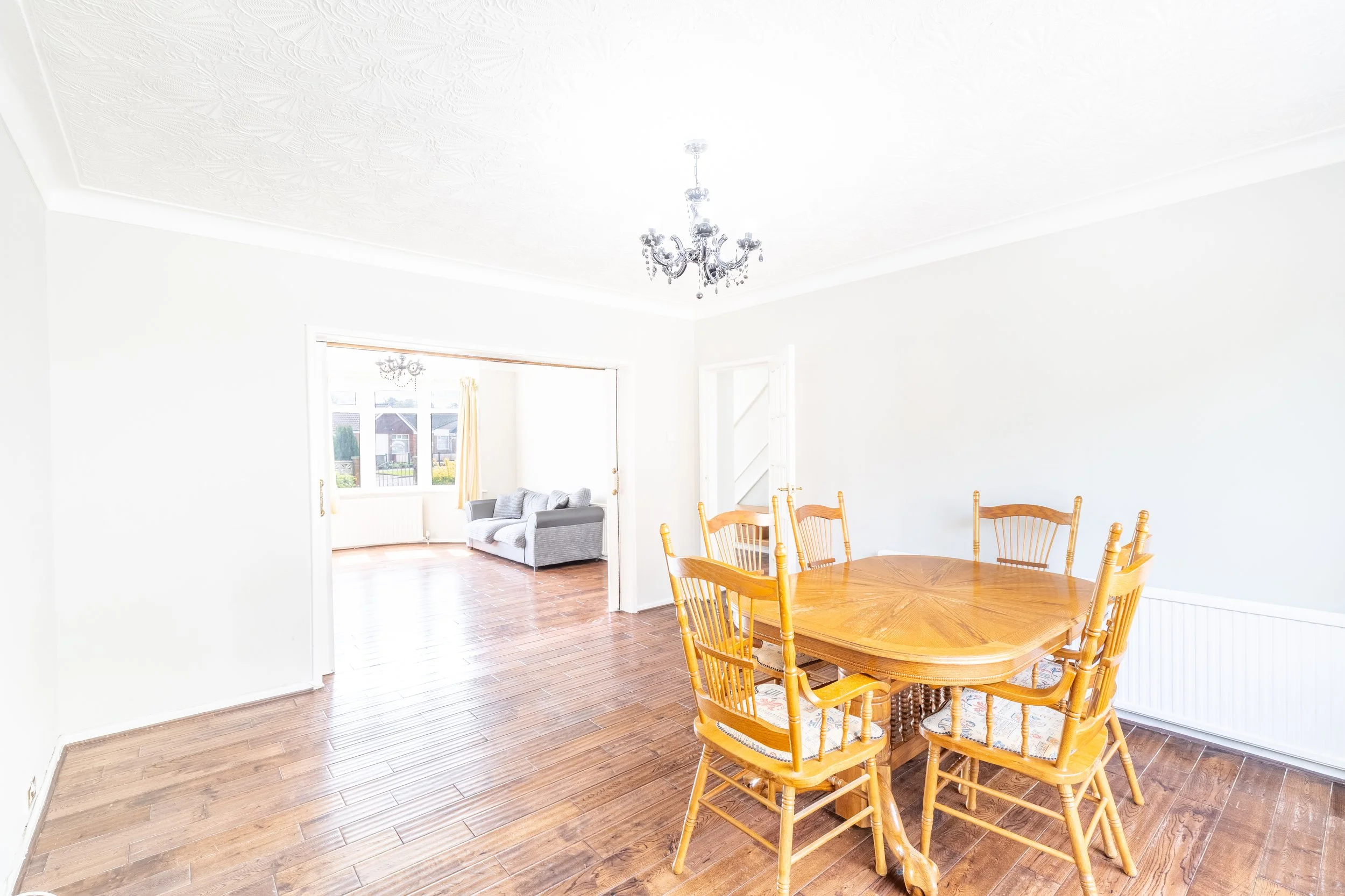 Bright dining room with wooden flooring and a dining table set. Adjacent open living area with a gray sofa and large window view.