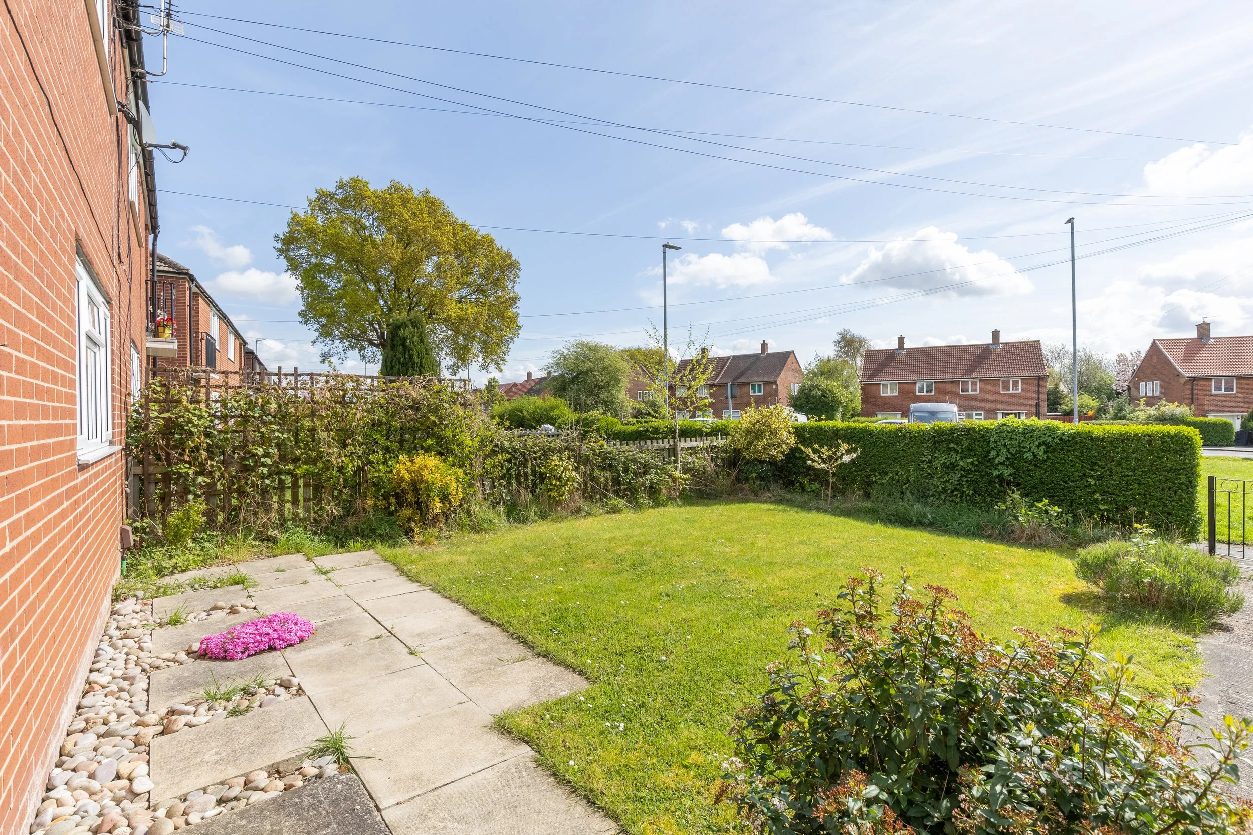 Residential backyard with lawn, pathway, hedges, and a tree, overlooking nearby houses under a blue sky.