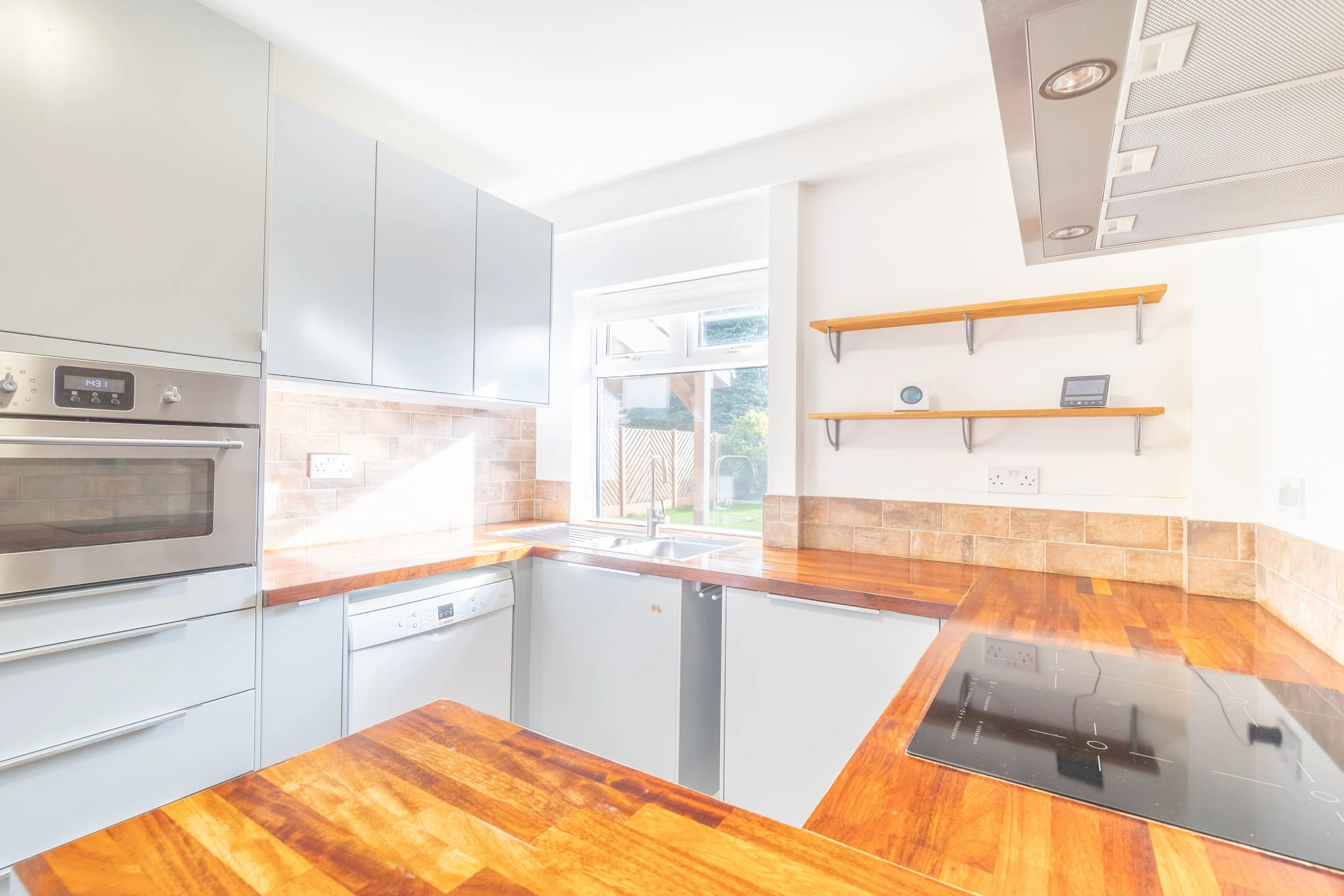 Modern kitchen with wooden countertops, stainless steel oven, white cabinets, tiled backsplash, induction cooktop, open shelves, and a window with garden view. Harrogate Kitchen Property Photo