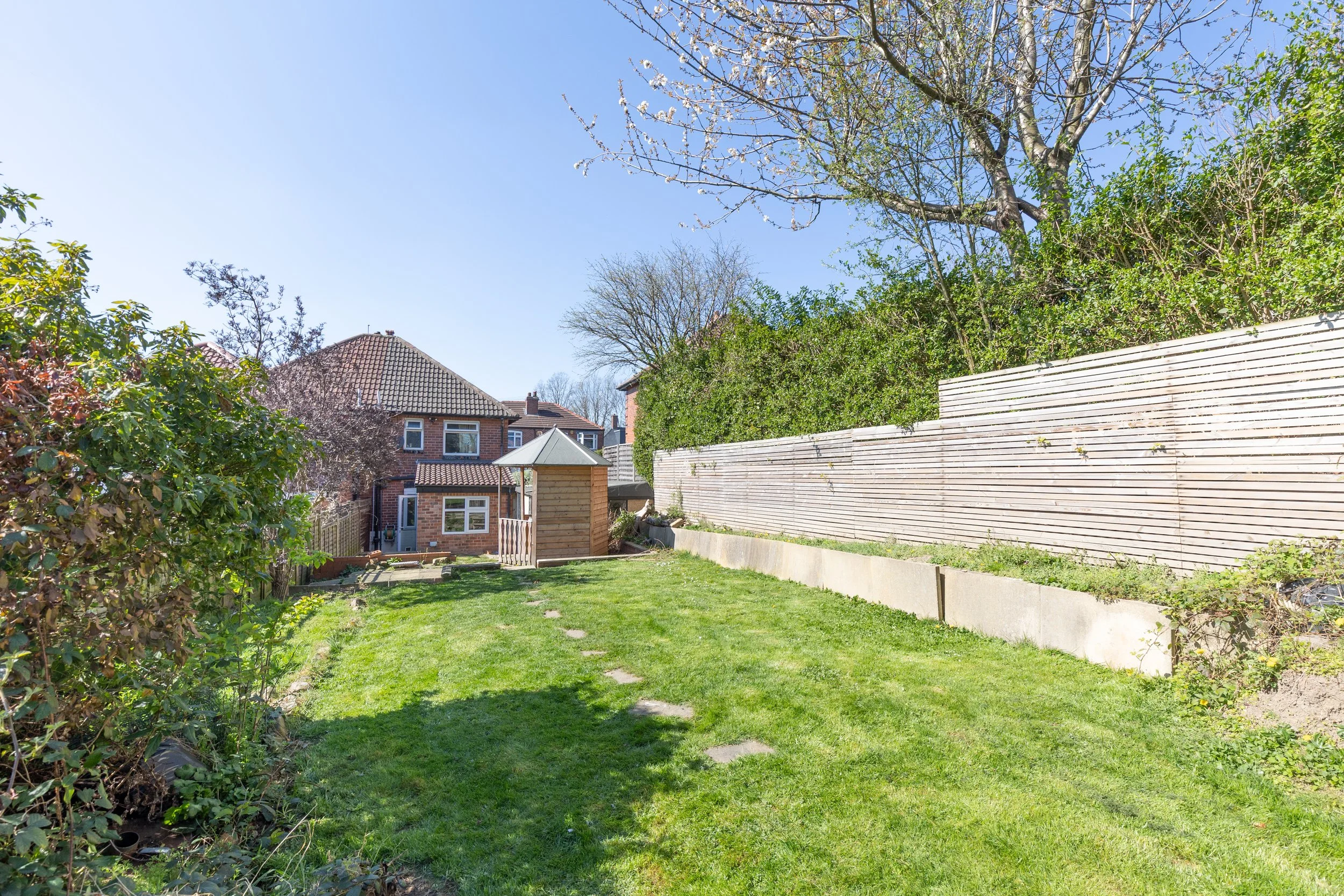 A backyard with a grassy lawn, wooden shed, and paved path leading to a brick house. The area is surrounded by trees and a wooden fence, with clear blue sky above.