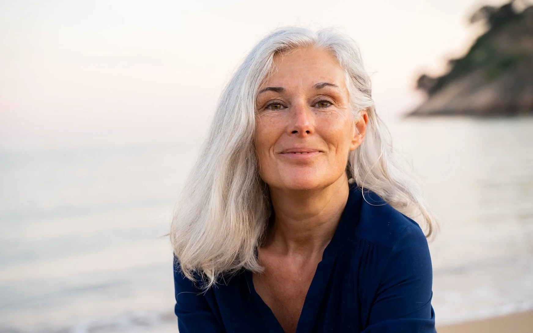 A close-up portrait of a mature woman with long gray hair, smiling gently, outdoors by the water during sunset or sunrise.