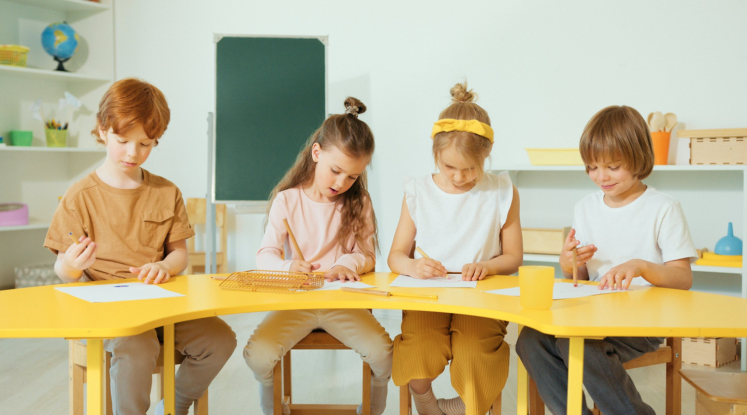 Four children sitting at a yellow table in a classroom, drawing on paper with pencils.