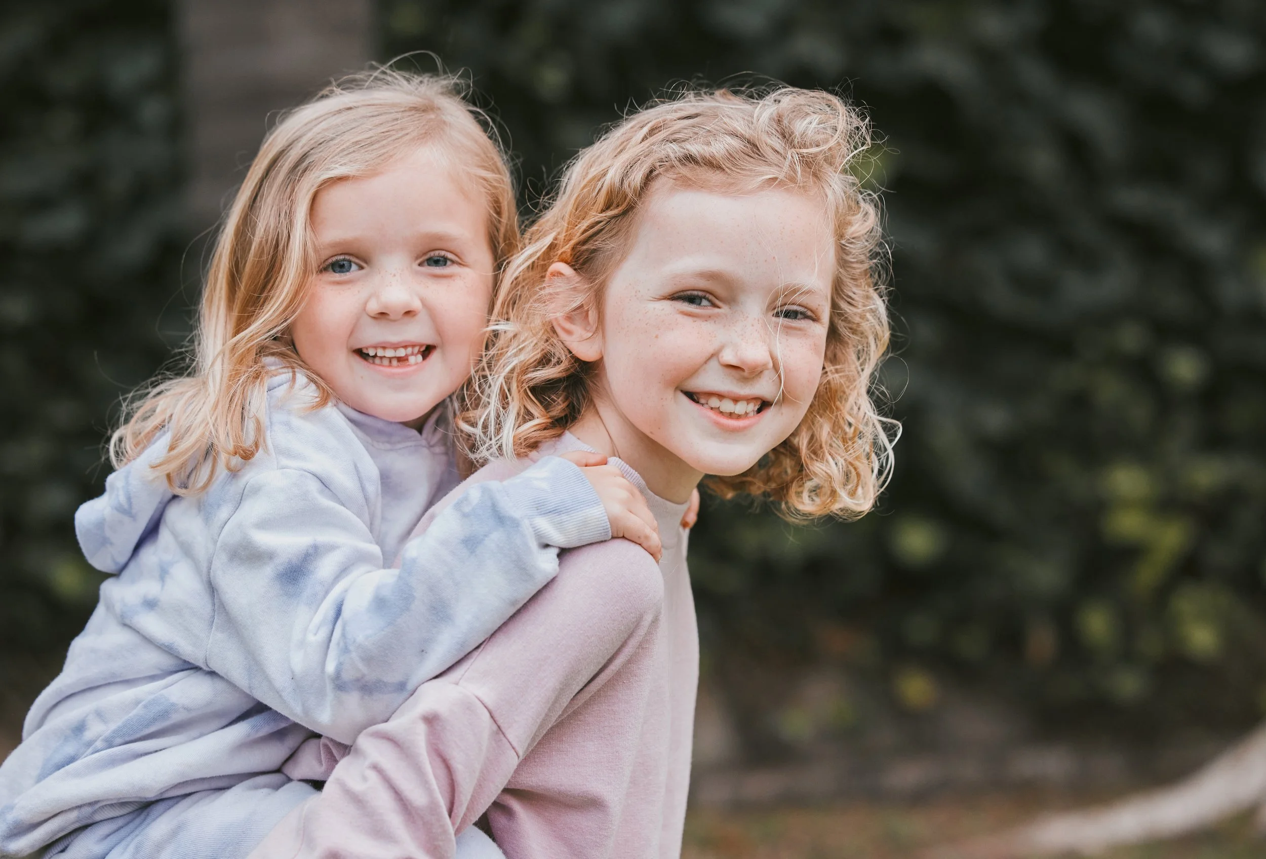 Two smiling young girls outdoors, girl with curly blonde hair giving piggyback ride, green foliage background.