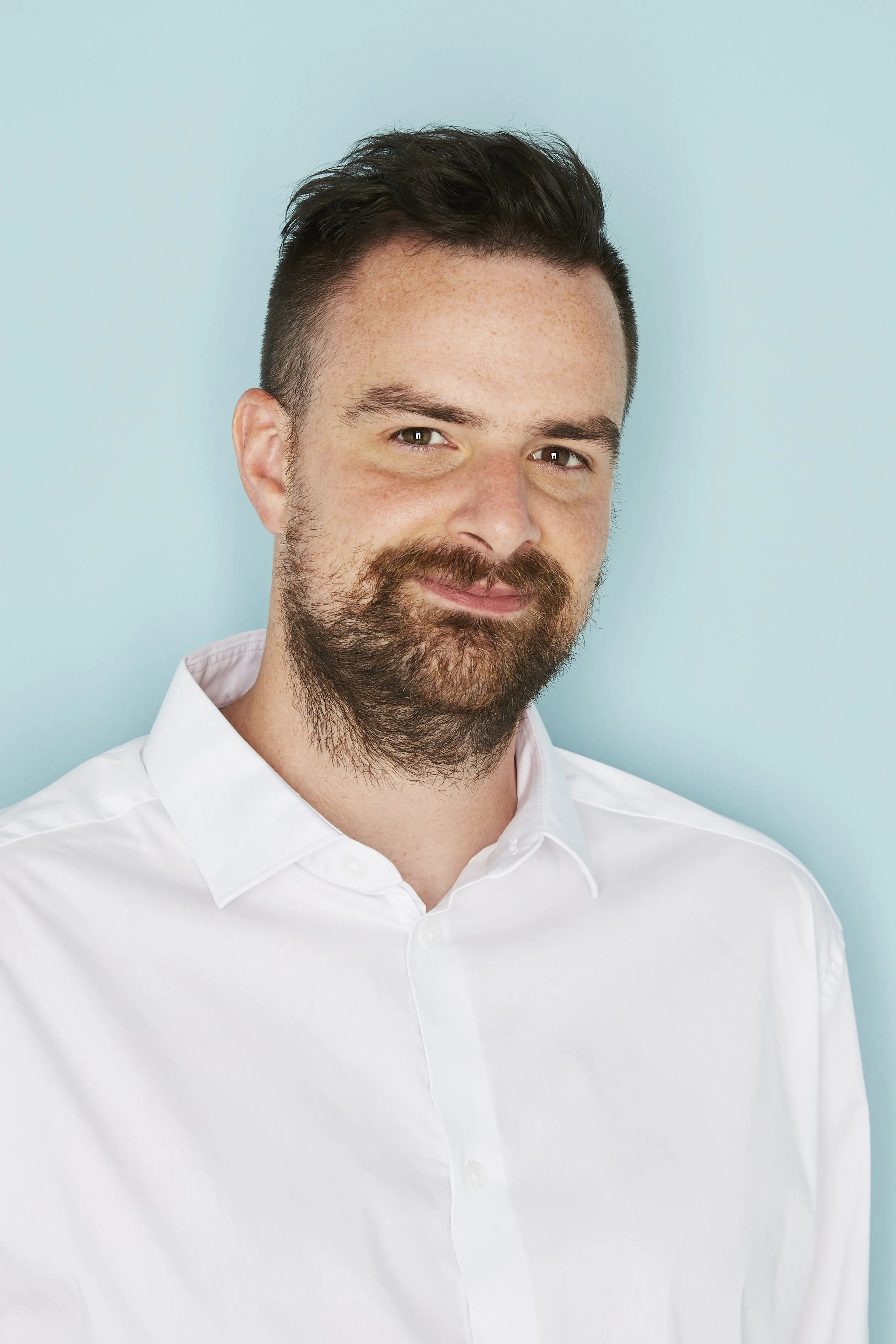 Greg Quee - Portrait of a smiling man with dark hair and a beard, wearing a white shirt, against a light blue background.