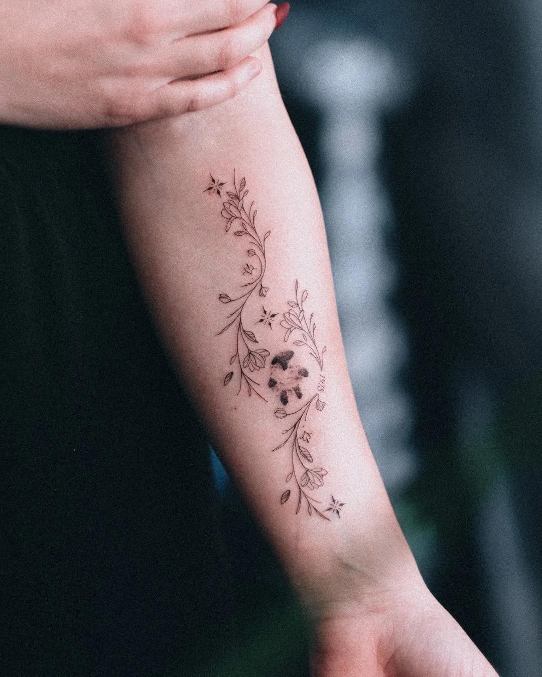 Fine line floral forearm tattoo with small flowers, leaves and date lettering by Leandro Correia in Zurich