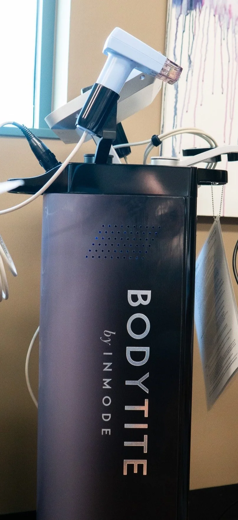 Medical device labeled 'BODYTITE' with a handpiece attached, in a clinical setting with a window and abstract artwork in the background.