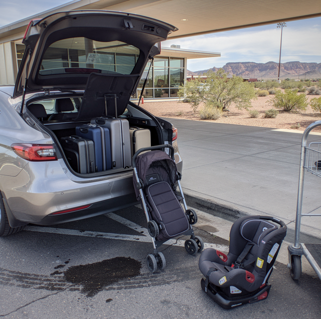 Silver car with open trunk loaded with luggage, stroller, and car seat in a desert parking lot.