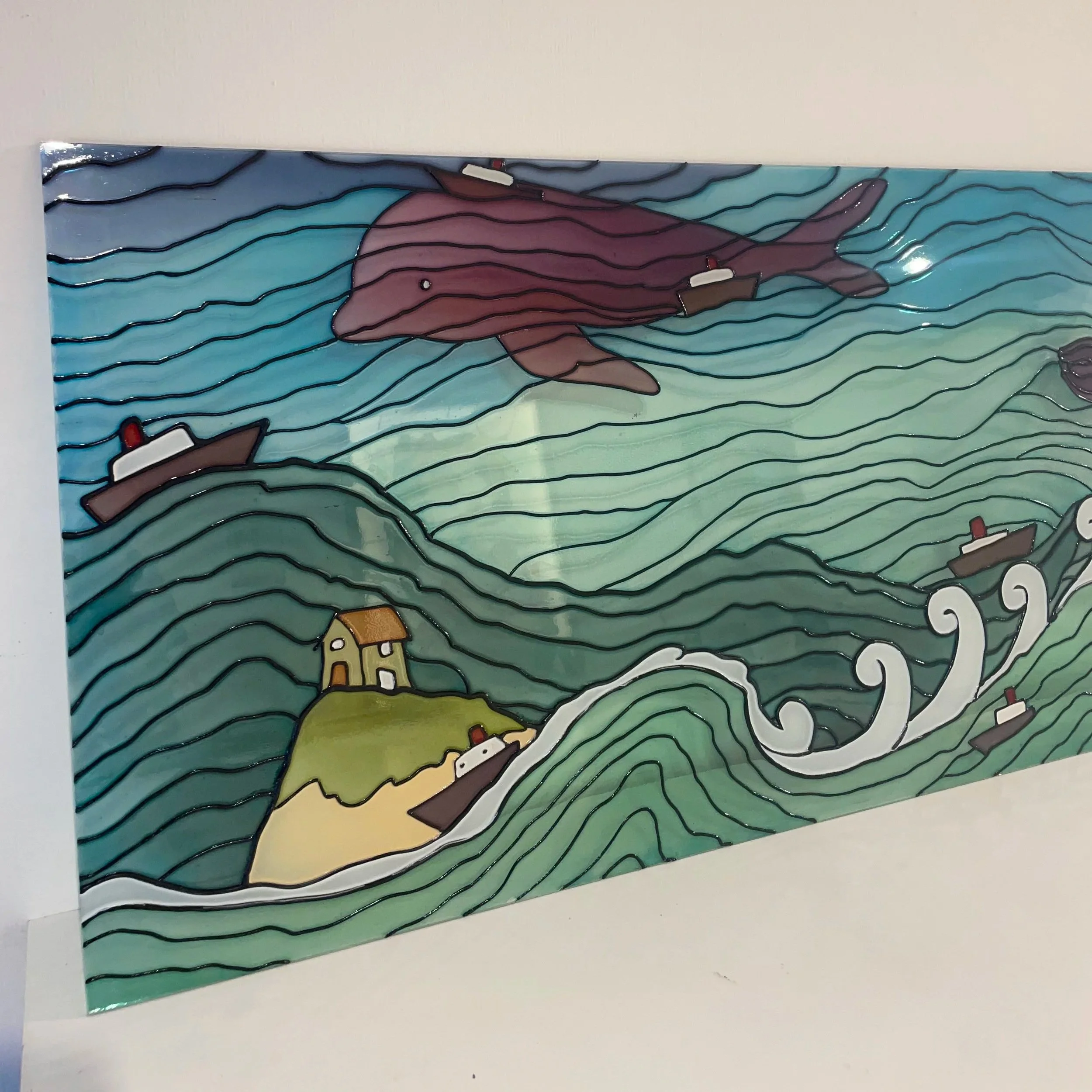 Colorful stained glass artwork depicting a large whale swimming among boats and land with houses, with stylized sea waves and a sky.