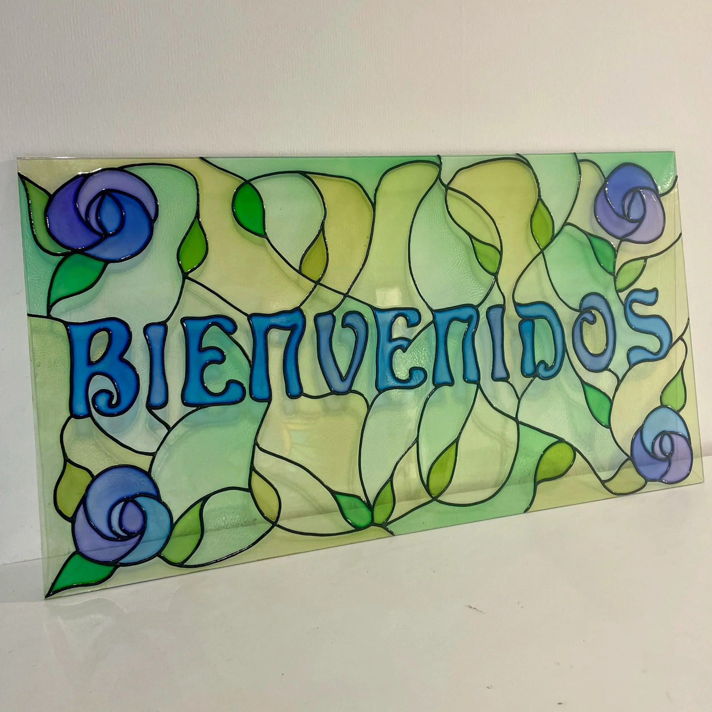 A stained glass sign with the word 'BIENVENIDOS' in blue letters, decorated with purple roses and green leaves.
