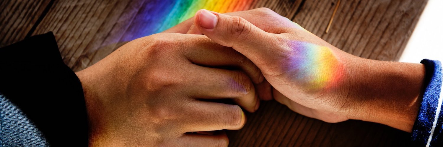 Two people are holding hands. Their hands rest on a wood table. A rainbow shines across them.