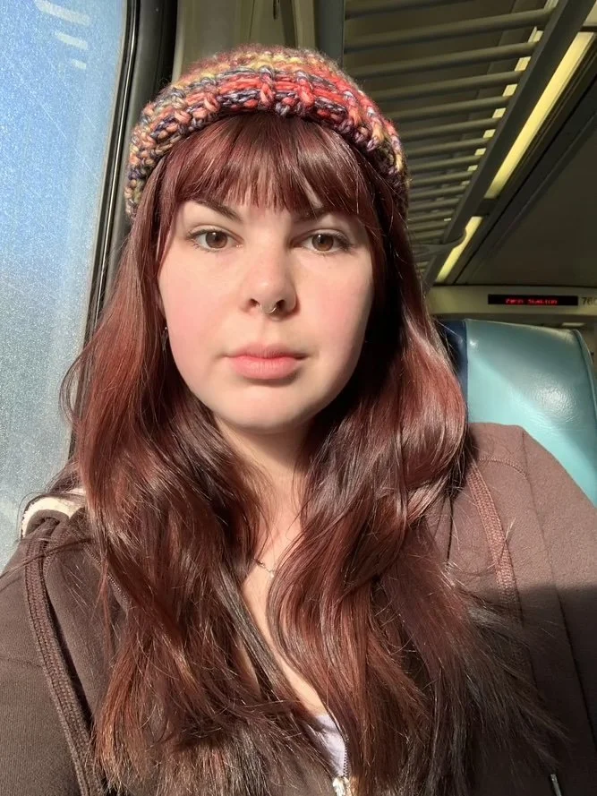 Headshot of Sofia Holden. Sofia has long, wavy, reddish-brown hair and is wearing a brown jacket and a knitted multicolor beanie. She is sitting by the window on a train.