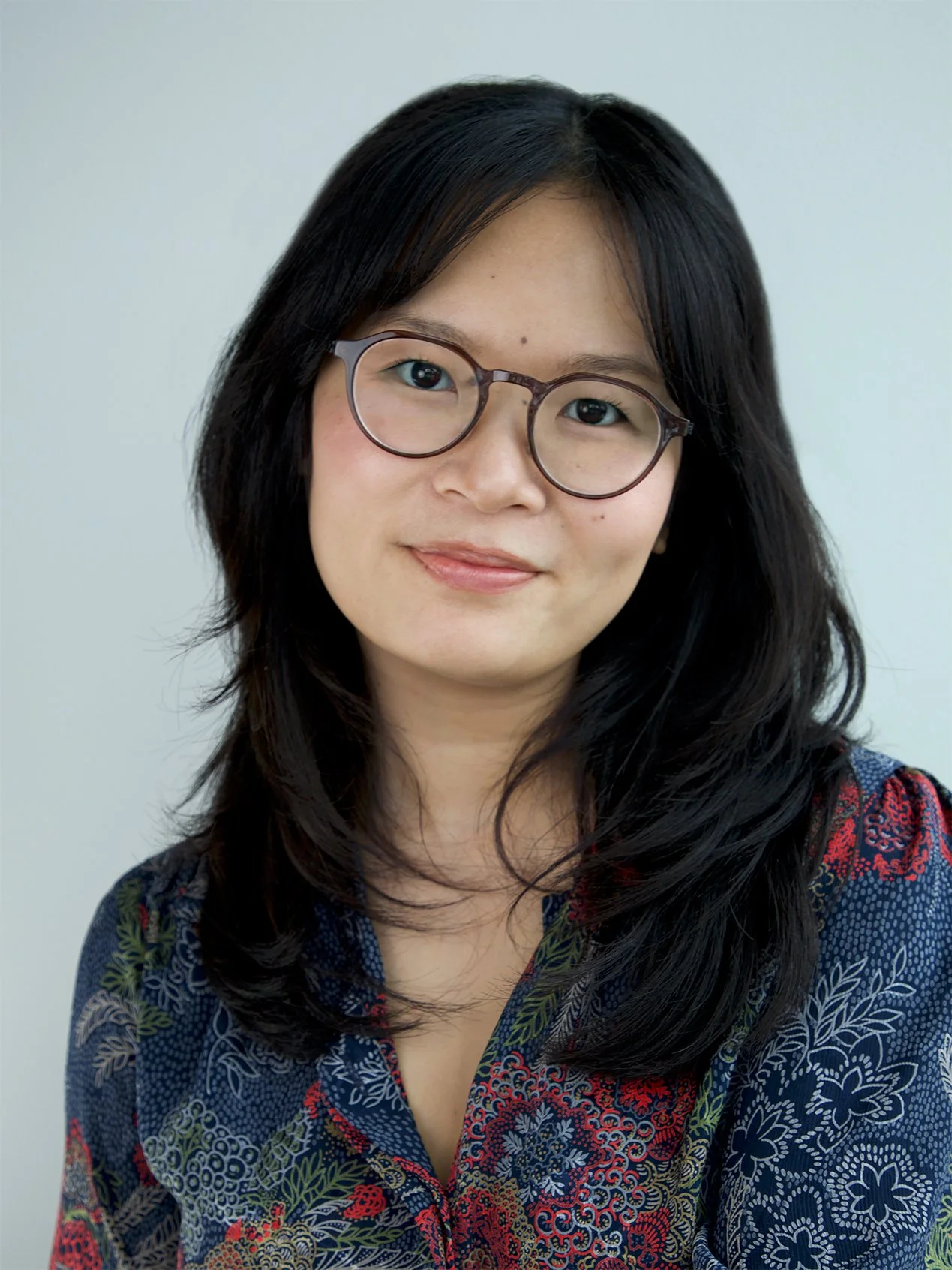Headshot of Ashley Wong in a beige blouse and yellow earrings in front a a white shelf with potted plants and a landscape painting.