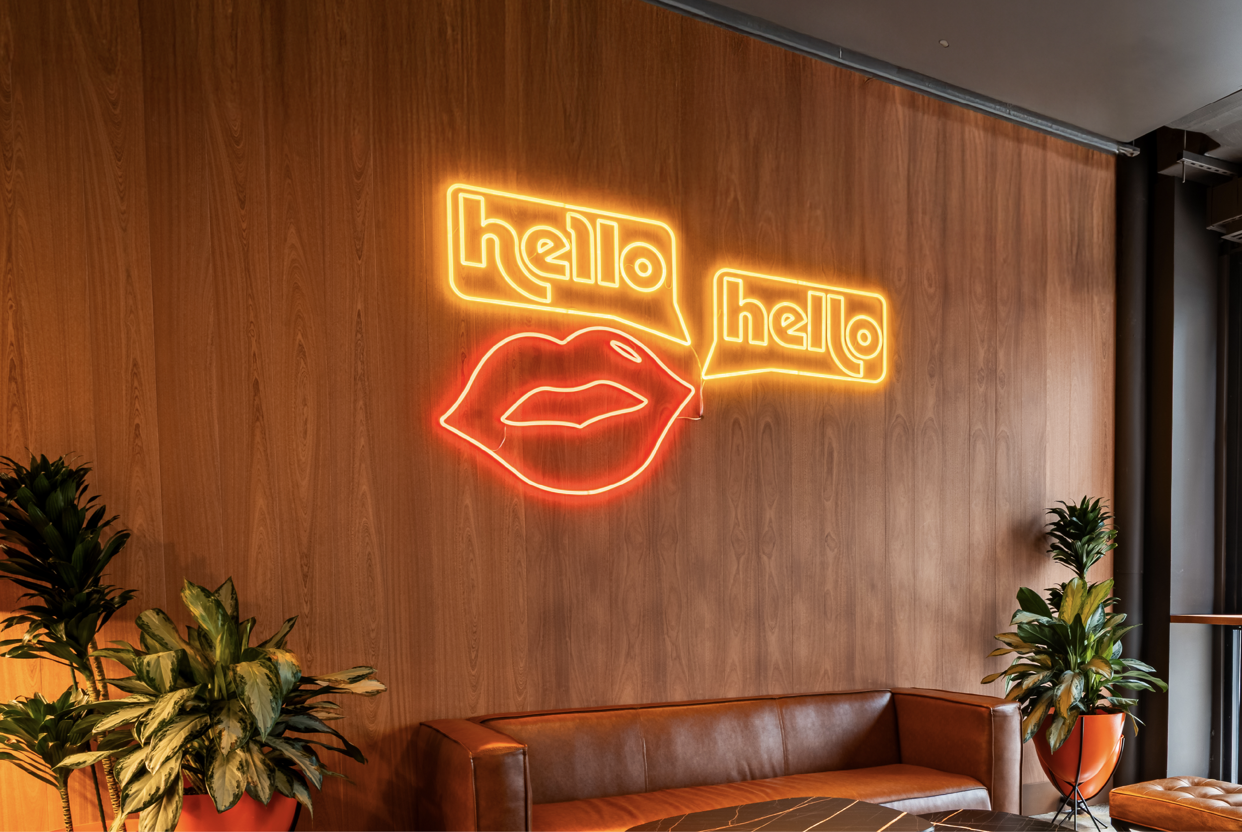 Neon sign on a wooden wall with the words 'hello hello' and lips graphic, near brown leather sofa and potted plants.