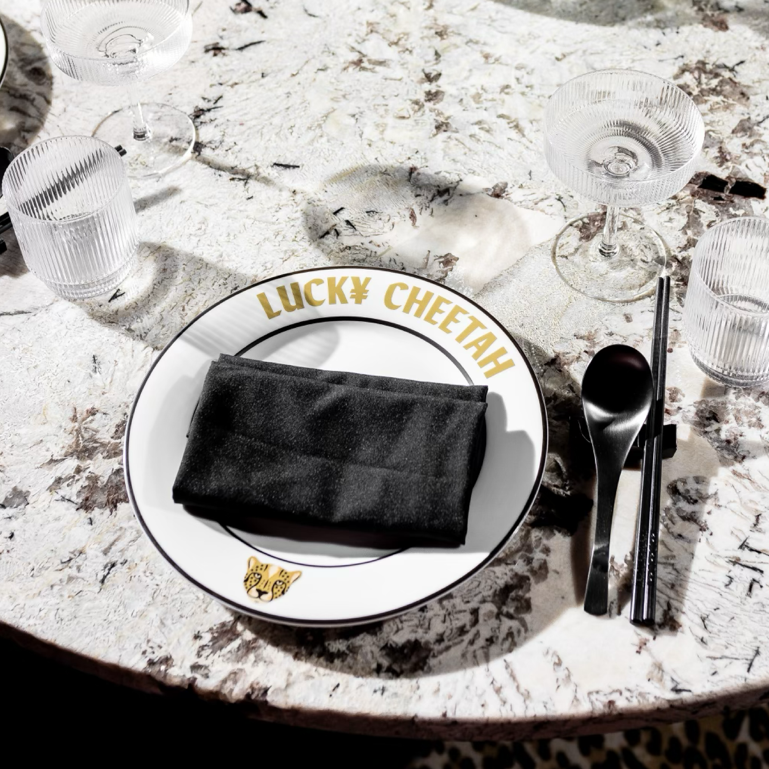 A place setting on a white, textured table with a Lucky CheeTah branded plate holding a black napkin. Surrounding the plate are four clear glasses, two with textured patterns and two with smooth surfaces. To the right of the plate are black utensils, a spoon, and a pair of chopsticks.